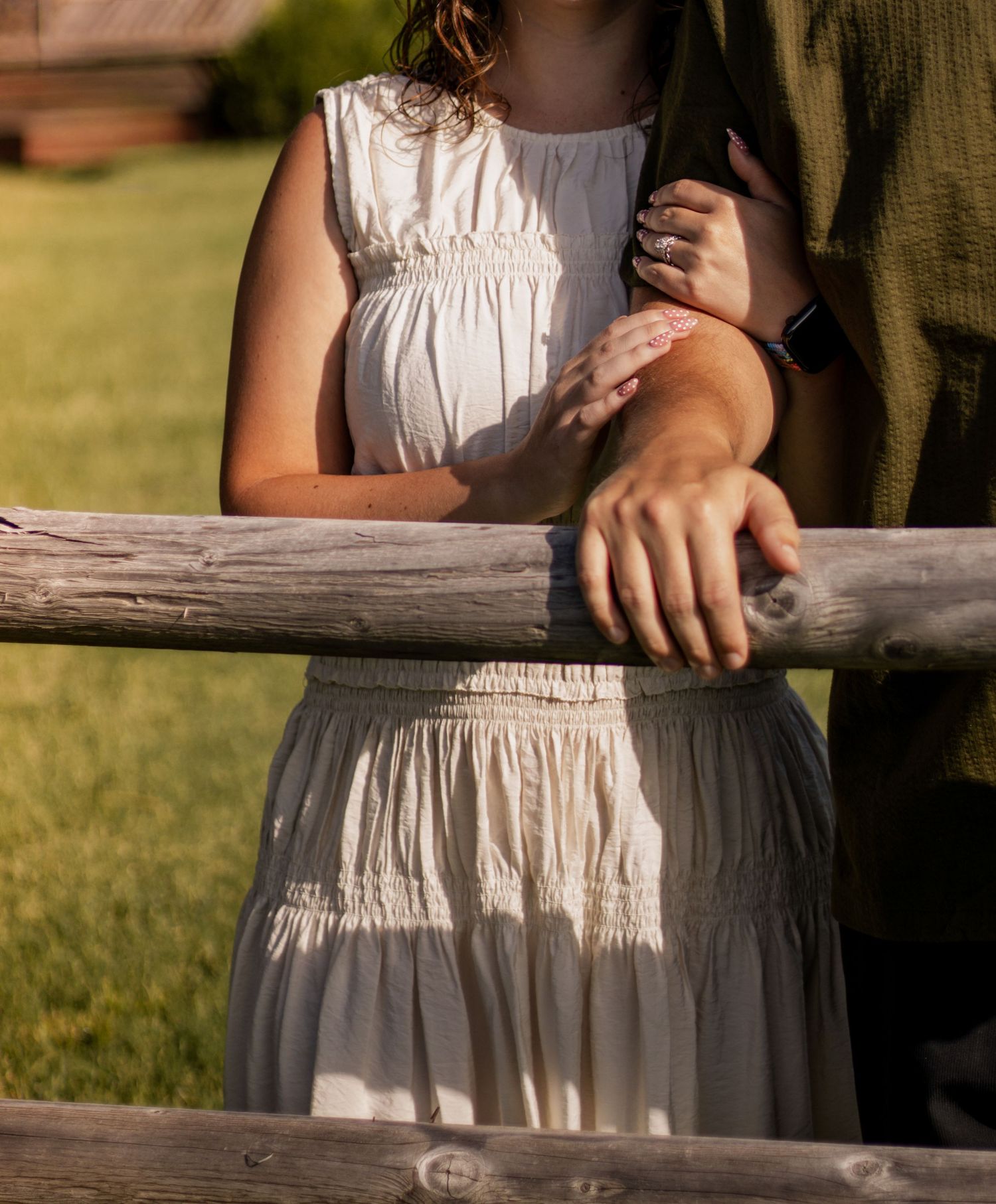 Couple standing together at a rustic wooden fence in a sunny green field, woman in white dress.