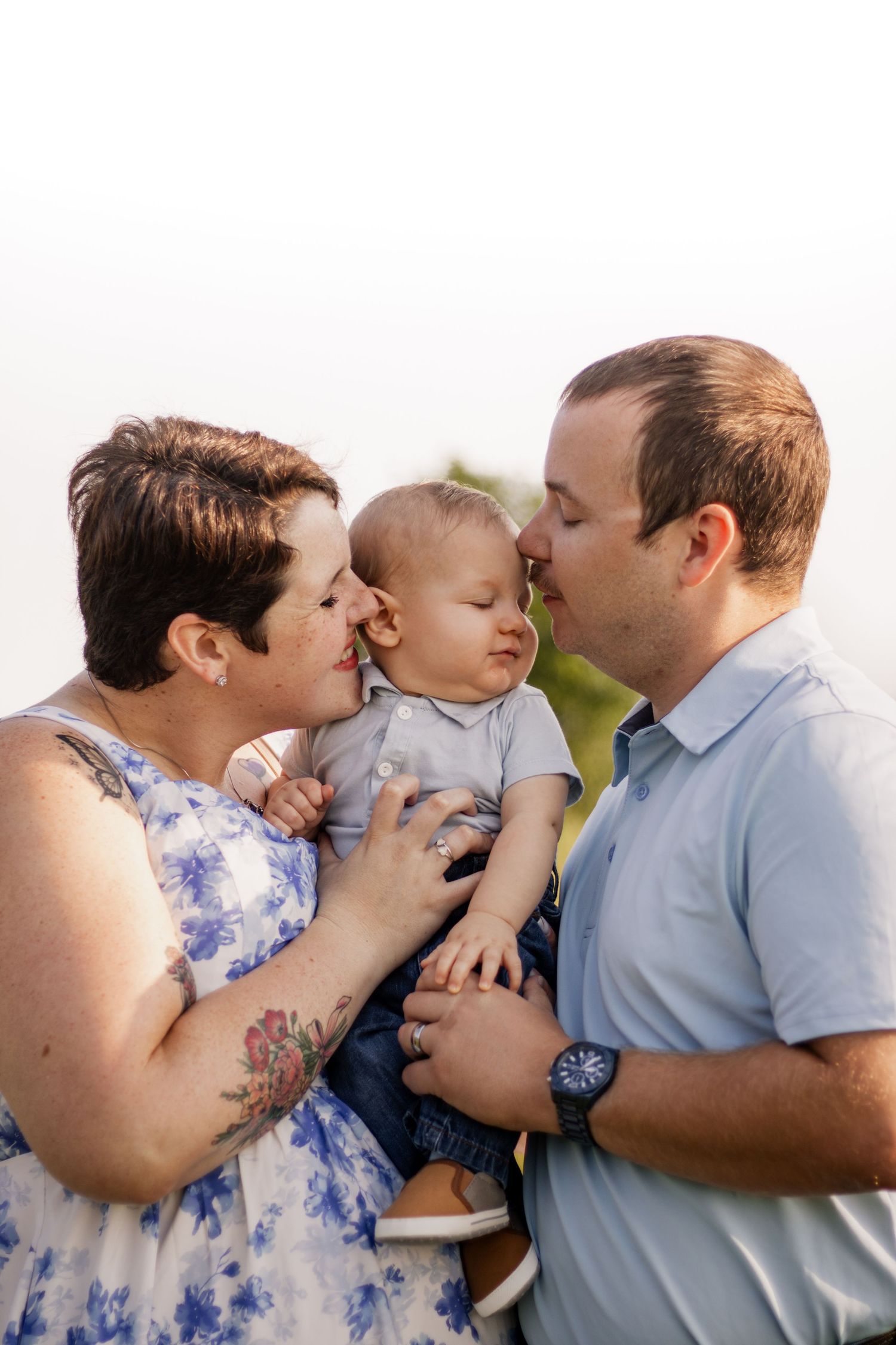 A couple kisses their smiling baby outdoors in a warm family portrait session.