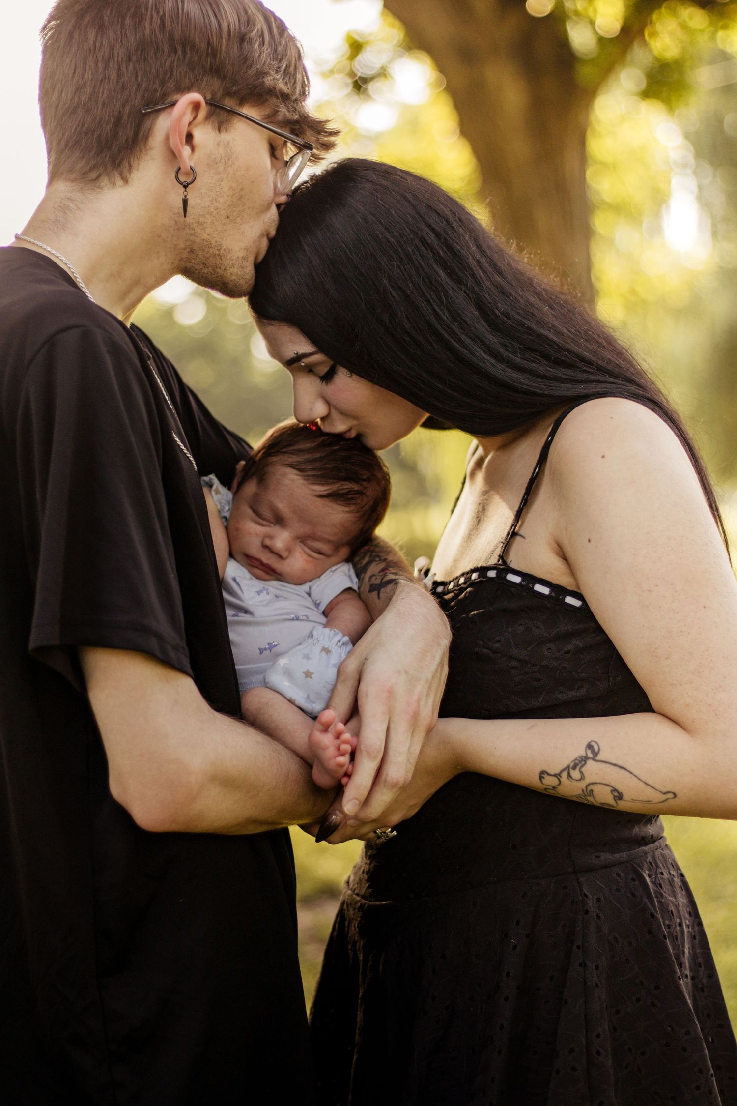 New parents tenderly kiss their sleeping newborn baby outdoors in a sunlit park, mother in black dress with tattoo.