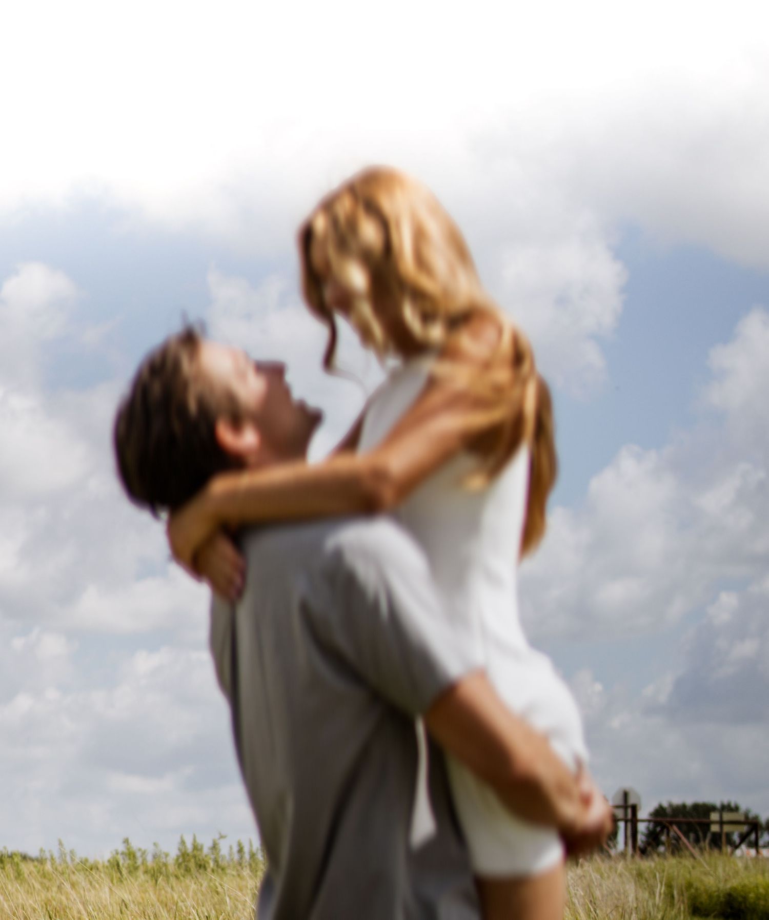A man lifts a woman in a romantic embrace in a sunny field with clouds in the background.