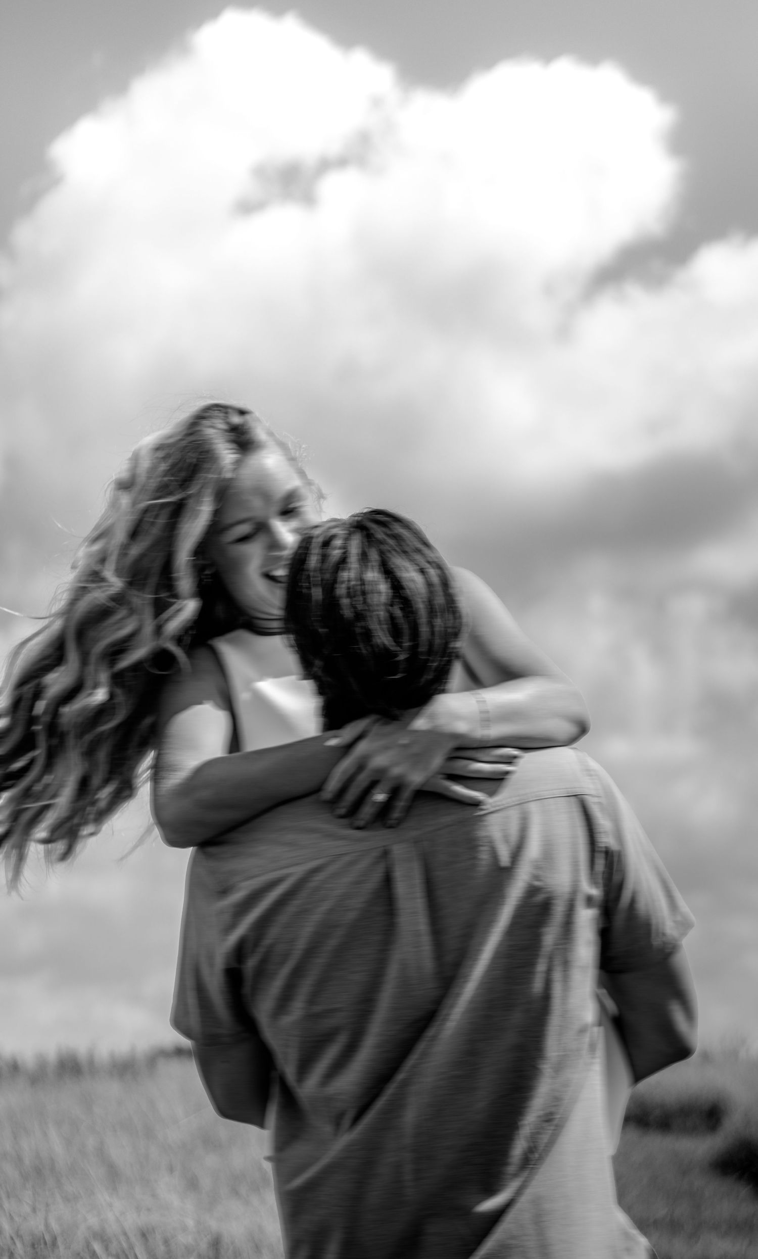 Man lifting and spinning woman in a joyful embrace under a cloudy sky in black and white photo.
