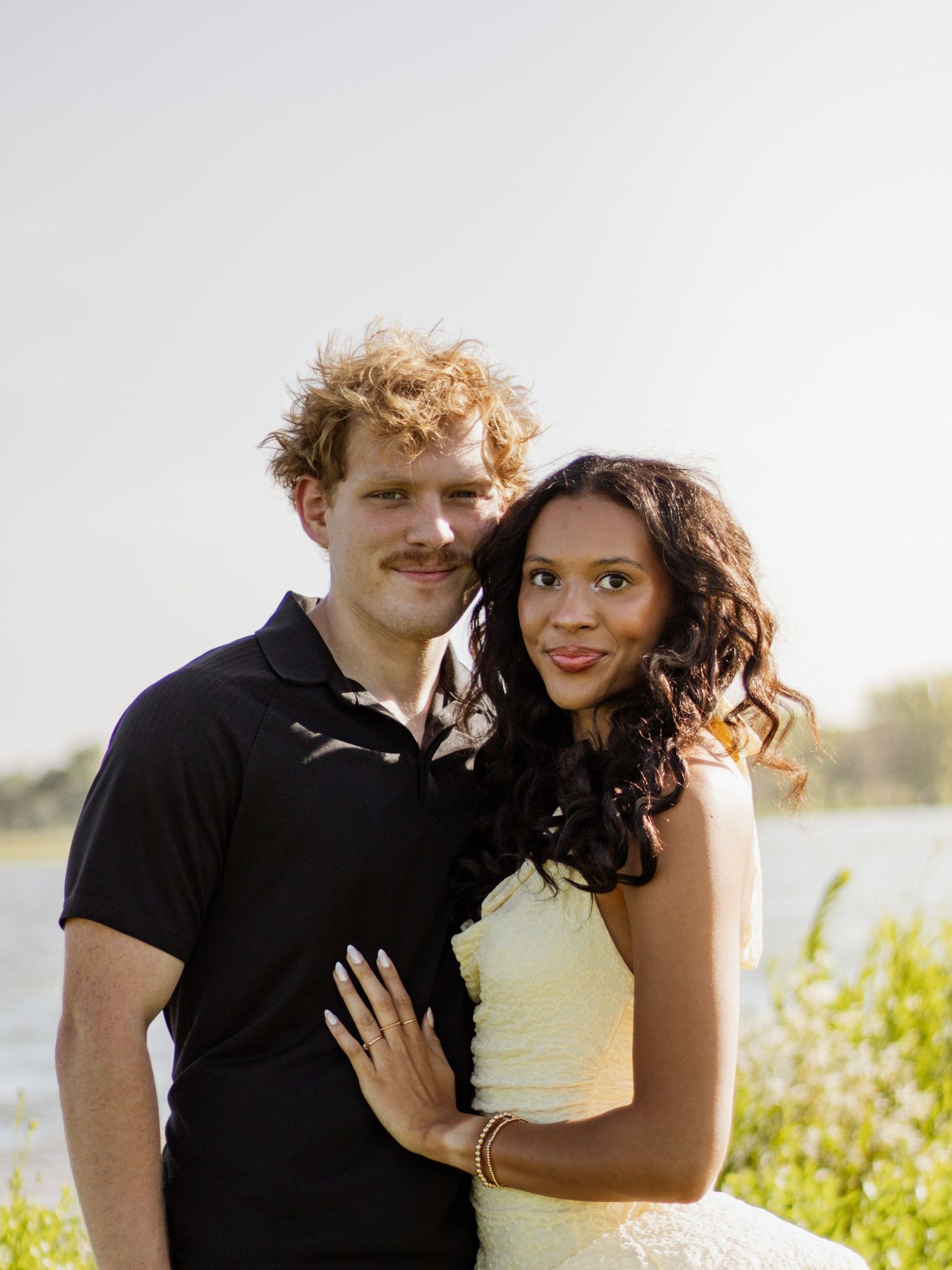 Smiling interracial couple posing outdoors near a lake, woman in white dress with gold bracelets, man in black polo shirt.