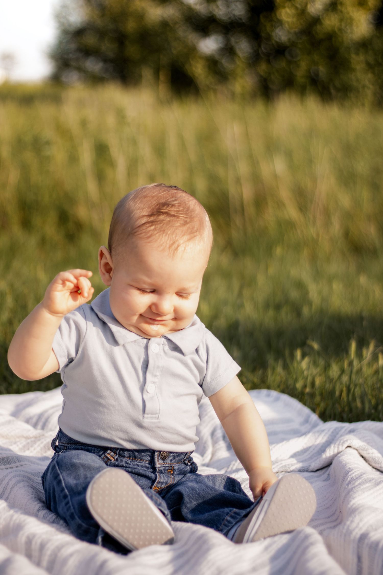 A happy baby boy in a gray shirt and jeans sits on a blanket outdoors in a sunny green field.