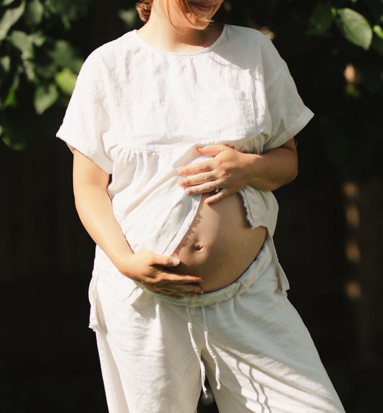 Pregnant woman in white outfit outdoors cradling her bare baby bump with both hands.
