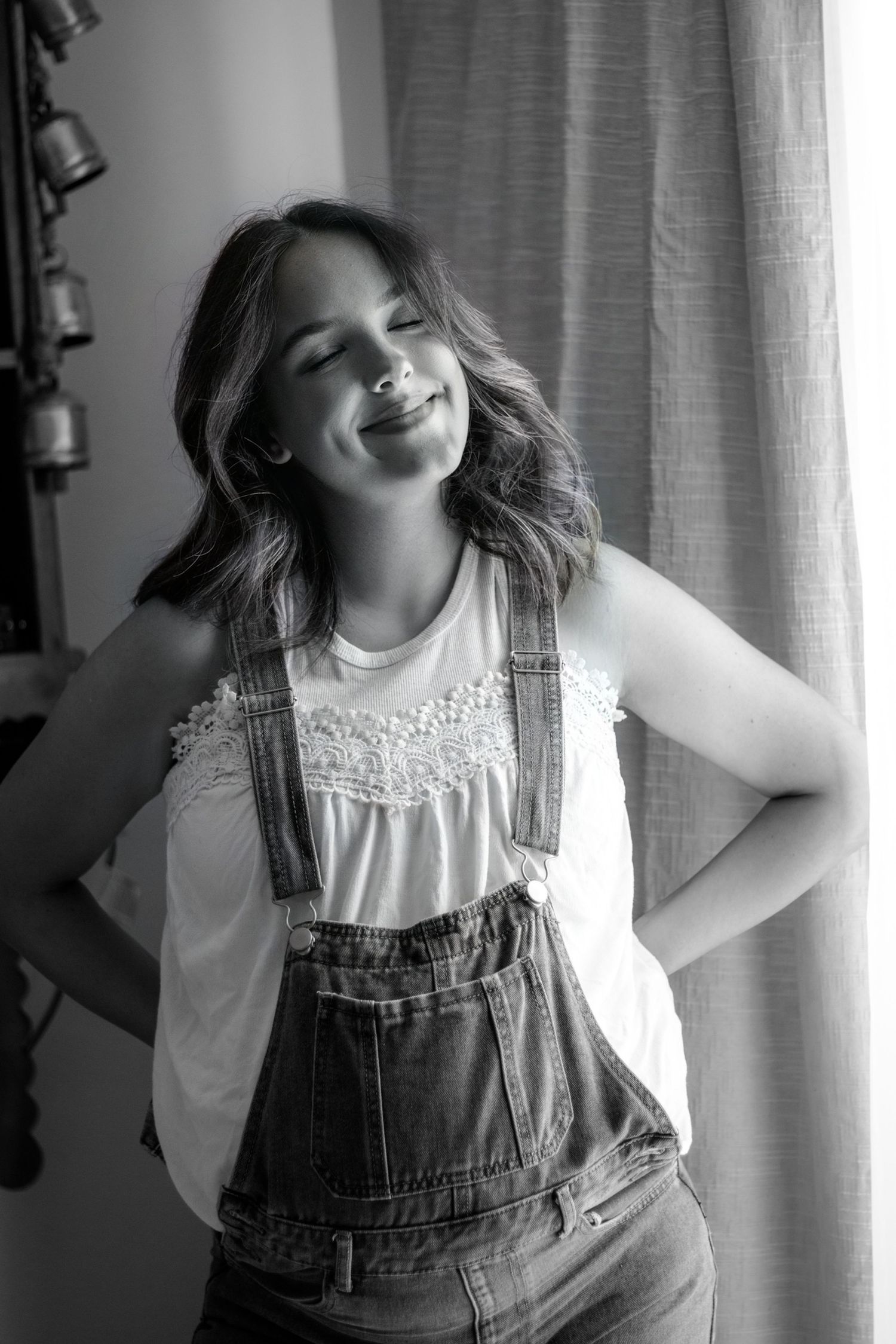 Black and white portrait of a smiling young woman in denim overalls and white top, posing with hands on hips.