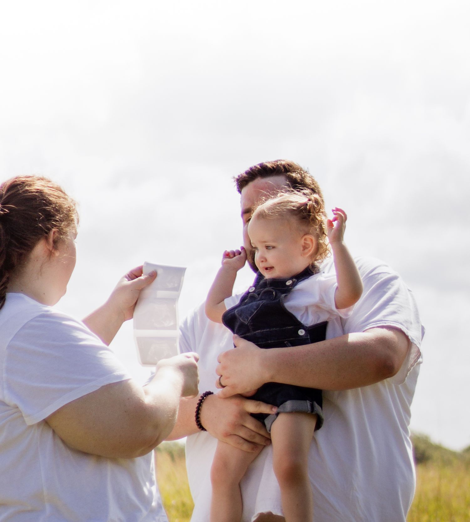 Family outdoors with man holding toddler while woman reads a document in a sunny field.