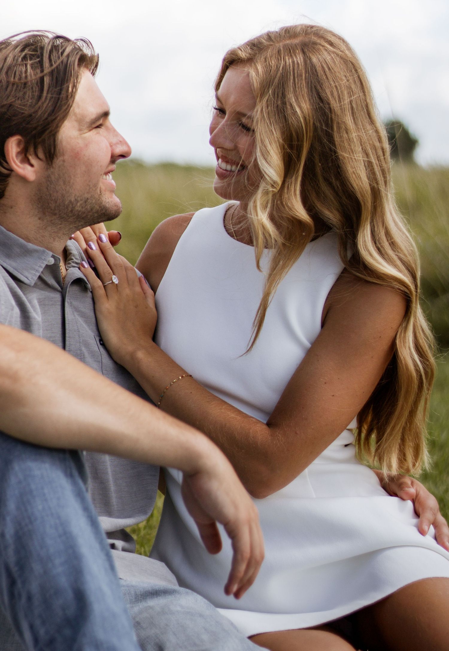 A smiling couple shares a romantic moment outdoors, woman in white dress showing engagement ring, sitting in grassy field.