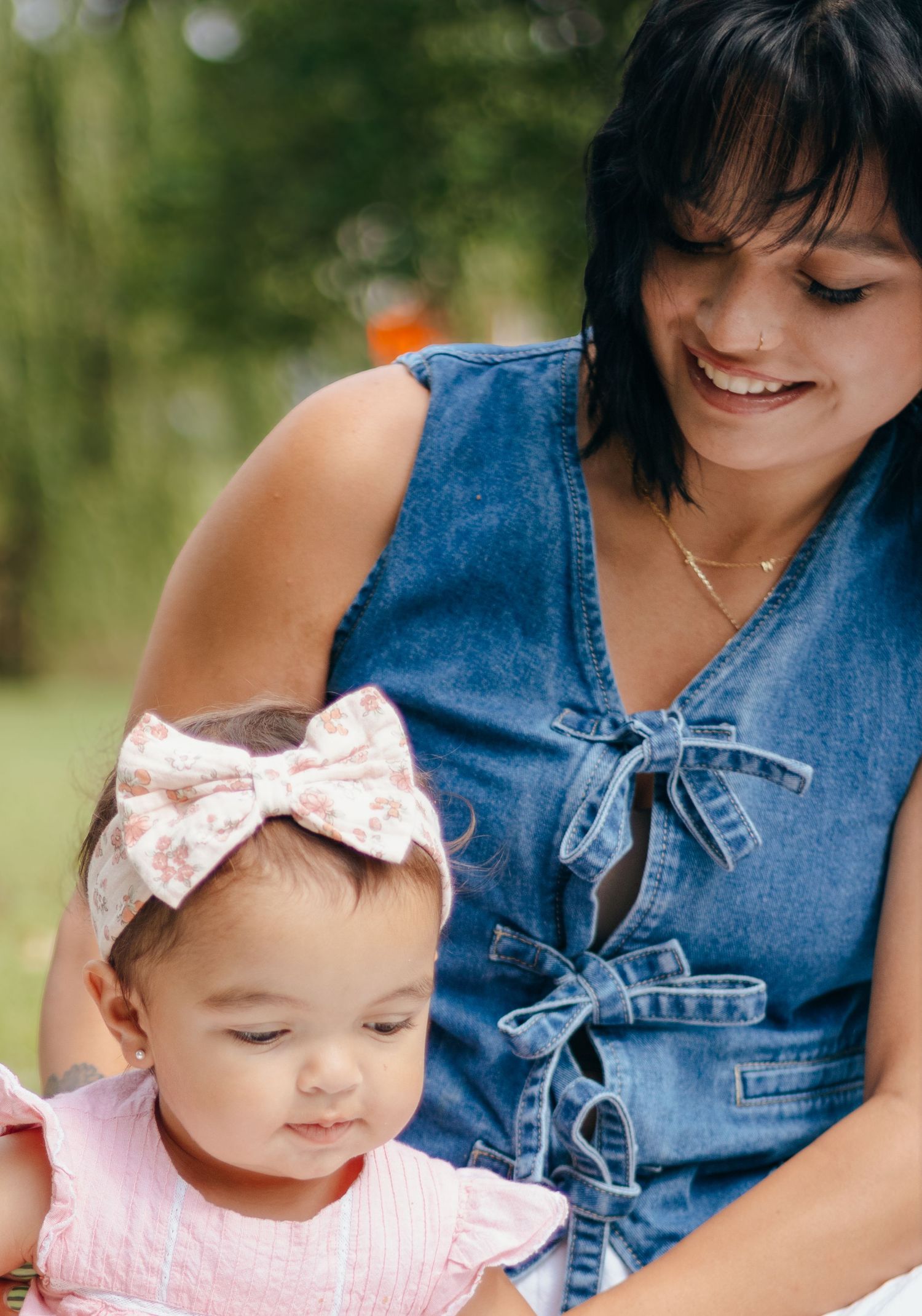 Smiling mother in denim dress looking at baby girl wearing pink bow headband outdoors.