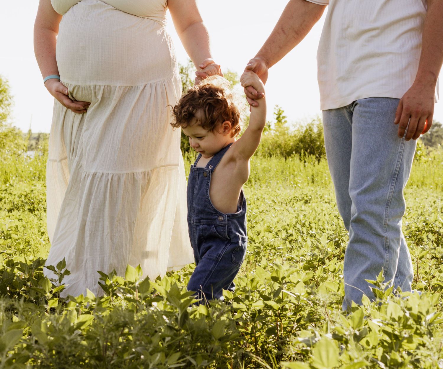 Pregnant woman in white dress and man hold toddler's hands walking in green field on sunny day.