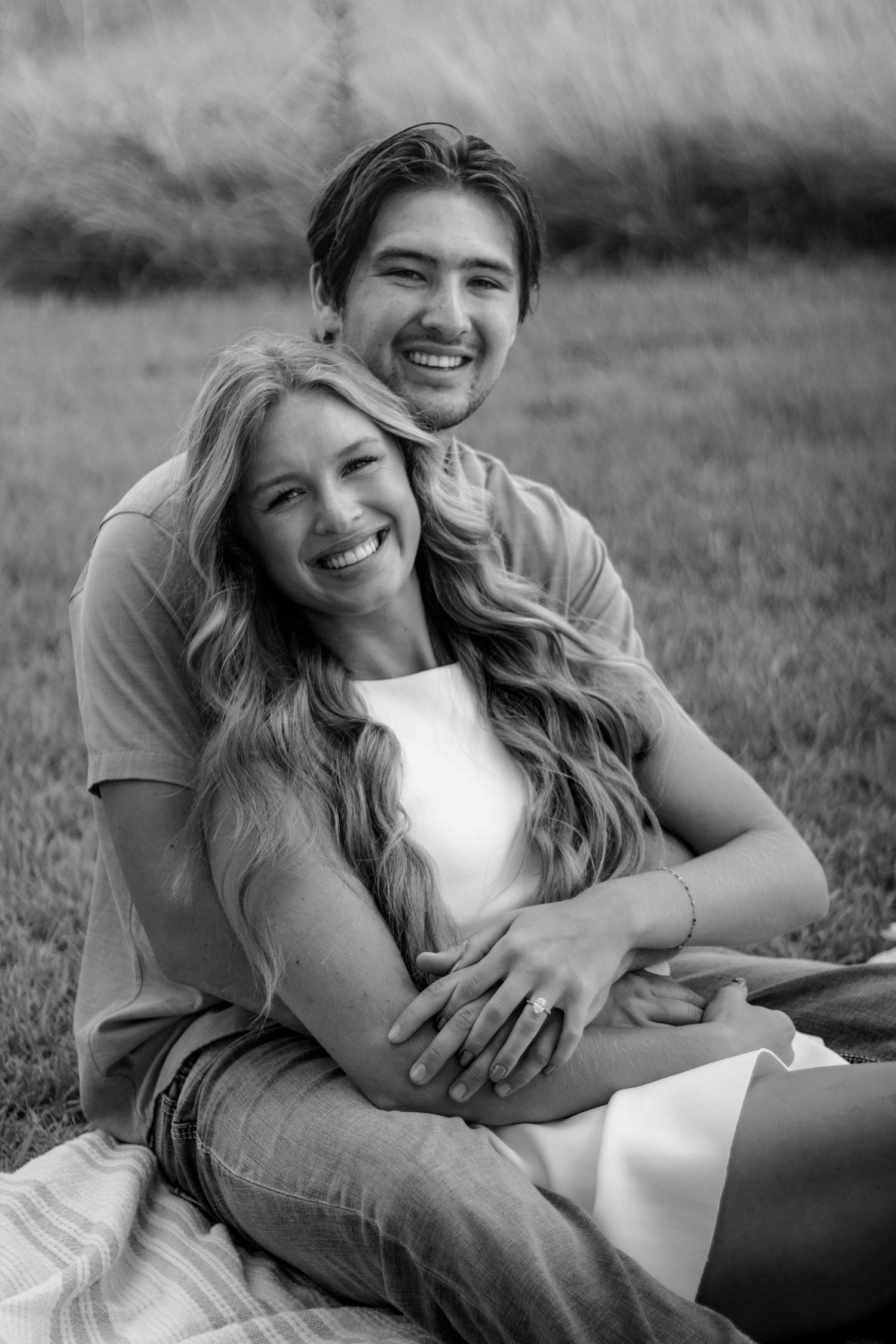 Black and white photo of a smiling couple sitting together on grass, man embracing woman from behind.
