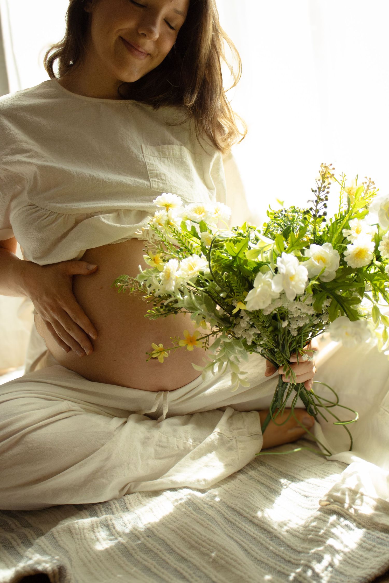 Pregnant woman with eyes closed smiling peacefully, holding wildflower bouquet while gently touching her bare baby bump.