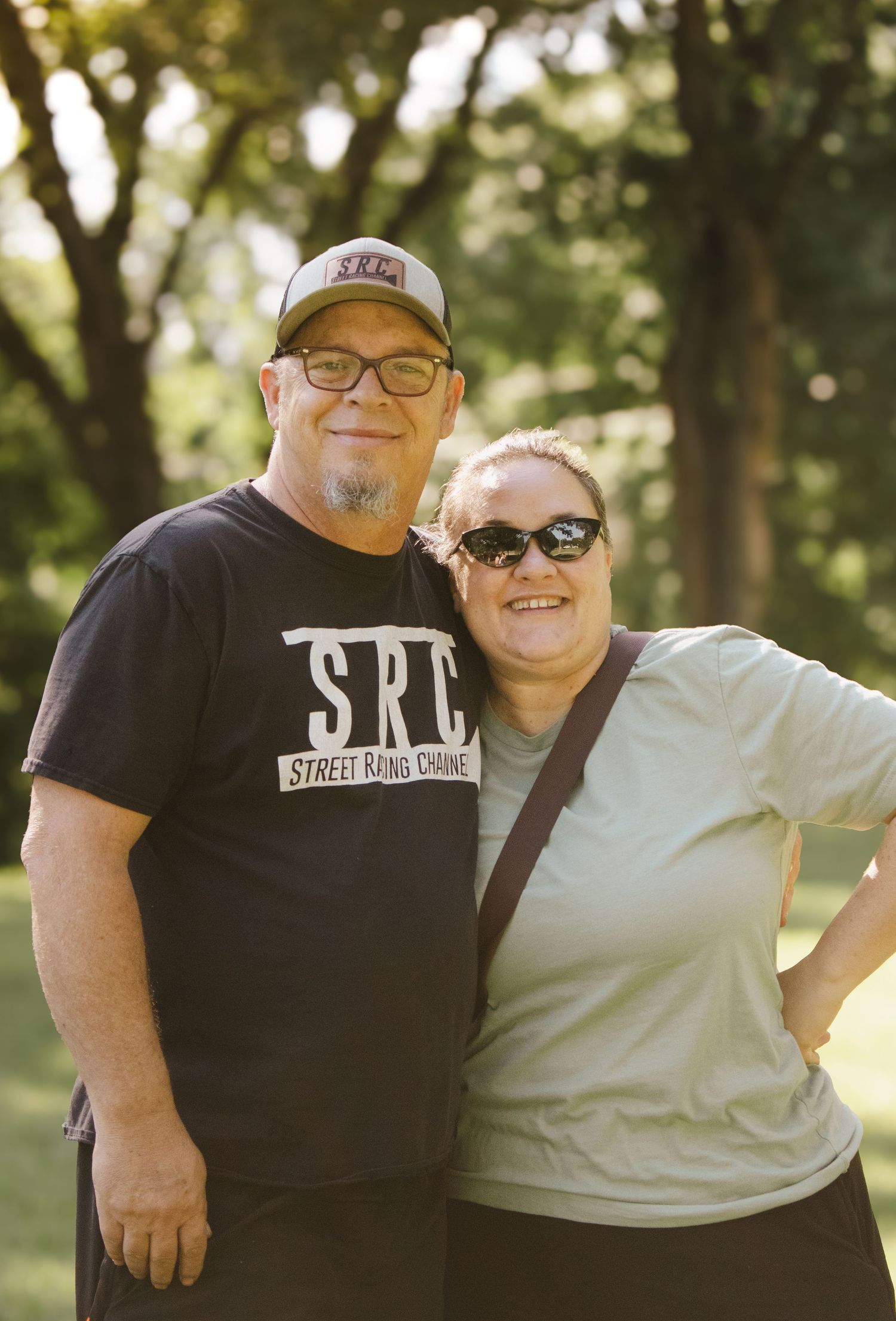 Happy couple smiling together outdoors in a sunny park, man in SRC black tee and woman in light green shirt.