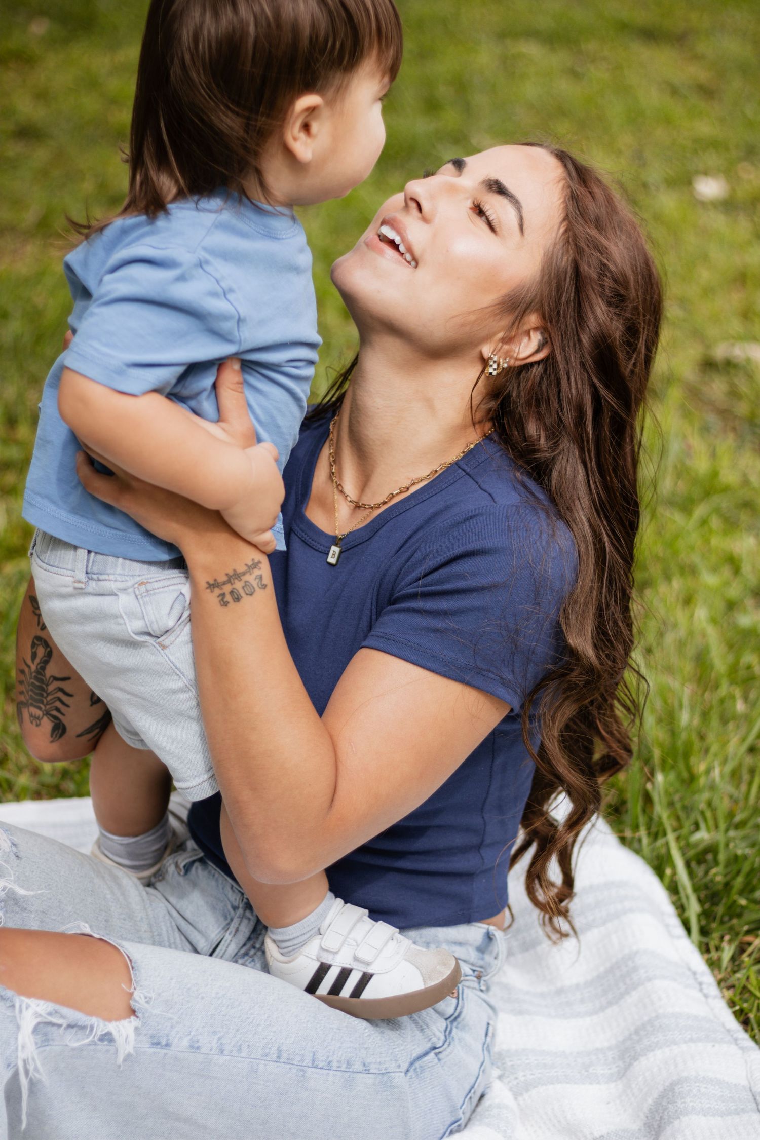 A young mother sits on a blanket in the grass, smiling up at her toddler son whom she holds in her arms.