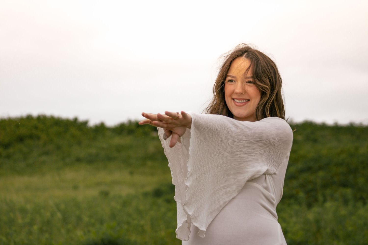 Smiling pregnant woman in white top extending arms forward in green field.