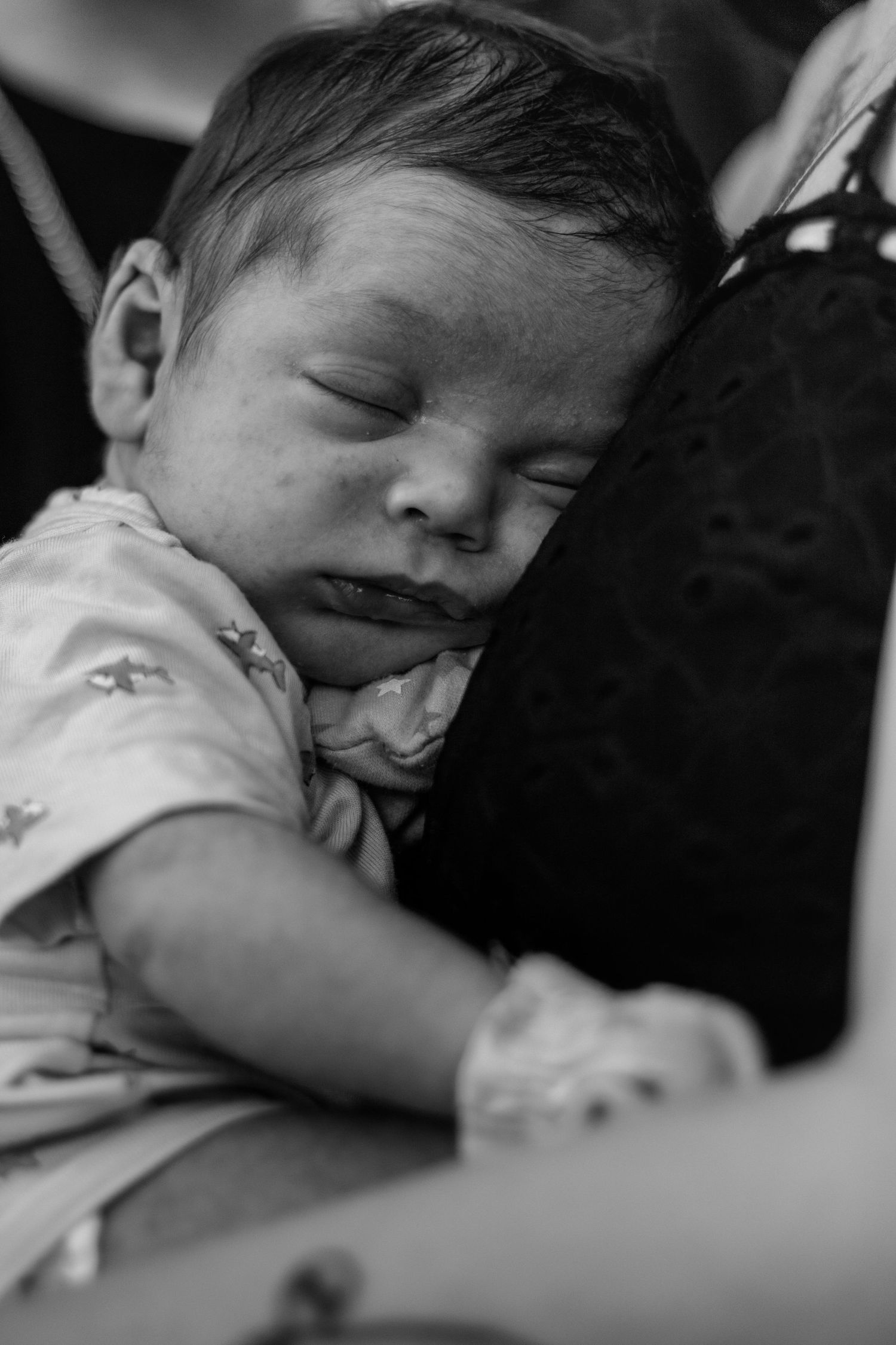 Black and white photo of a sleeping newborn baby resting peacefully against a person, wearing a patterned onesie.