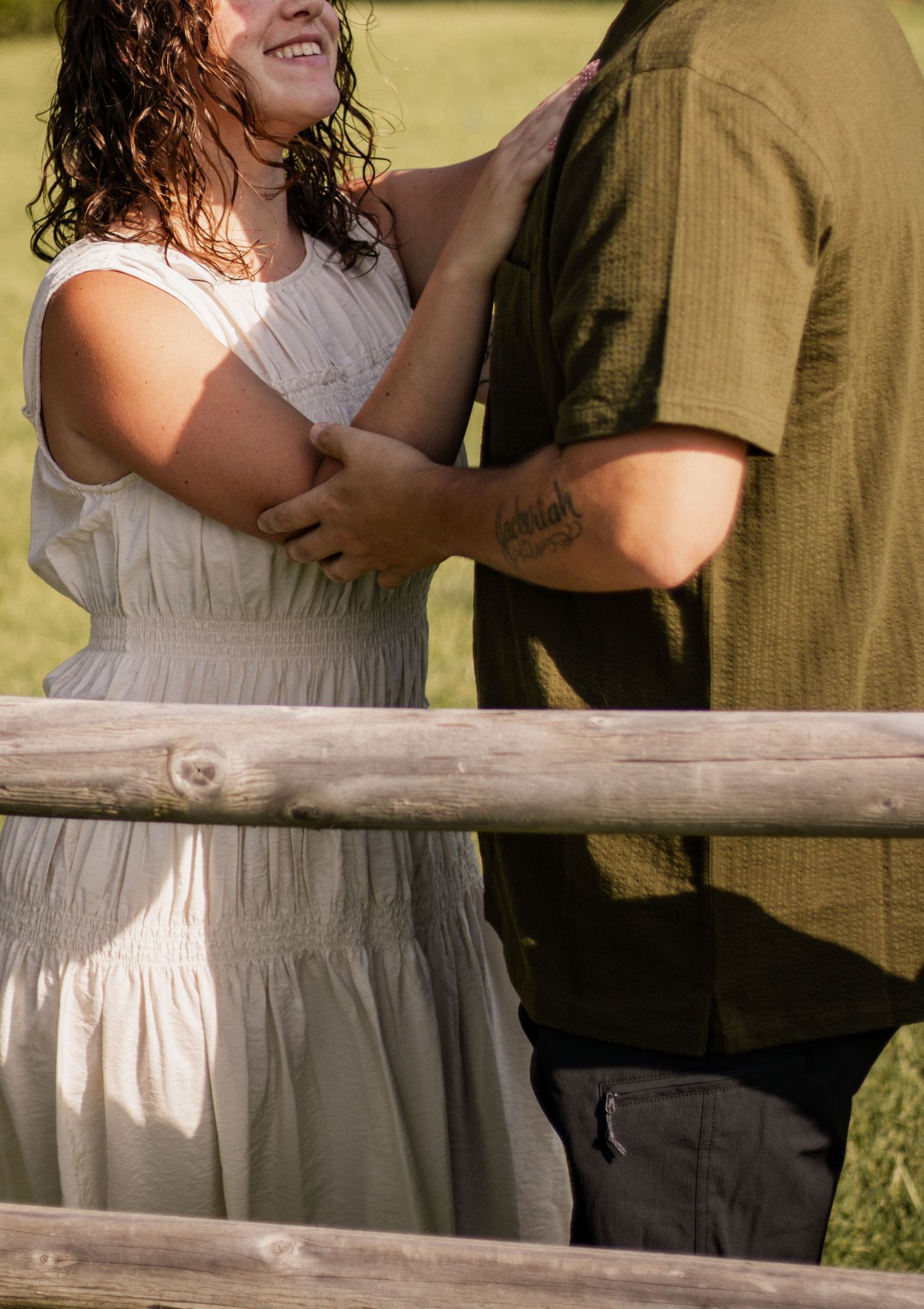 Smiling woman in white dress embraces man in green shirt by rustic wooden fence in sunny outdoor setting.