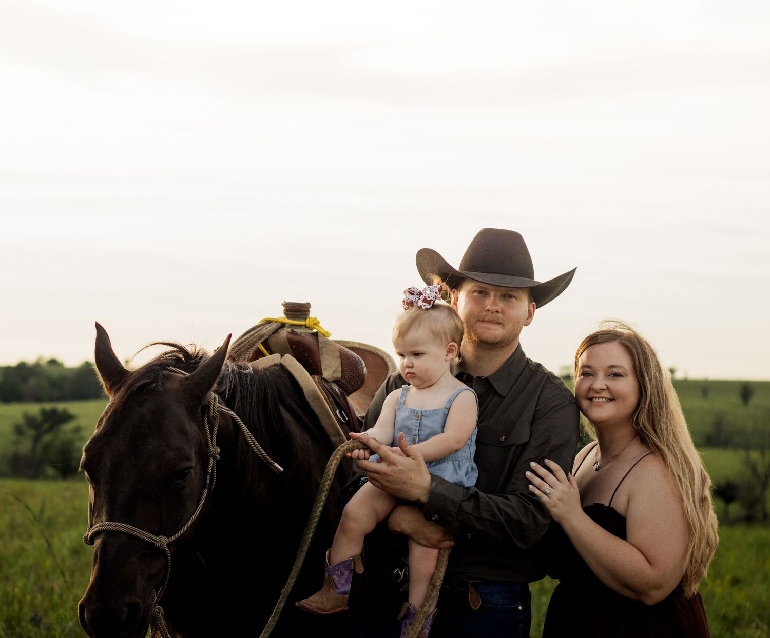 Young couple with baby and black horse in green field, man in cowboy hat, country family photo outdoors.