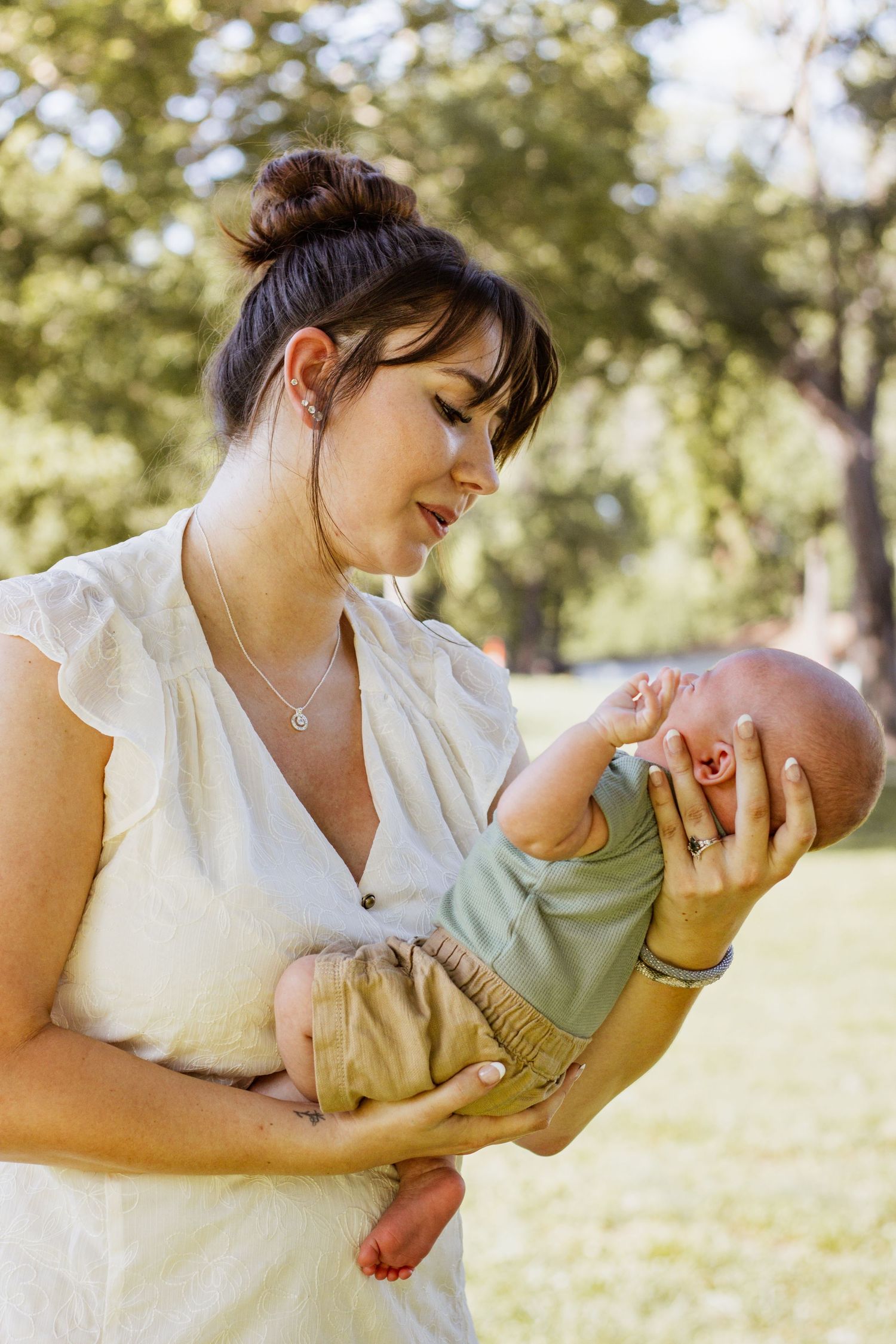 A young mother in a white dress tenderly holds her newborn baby outdoors in a sunny green park.
