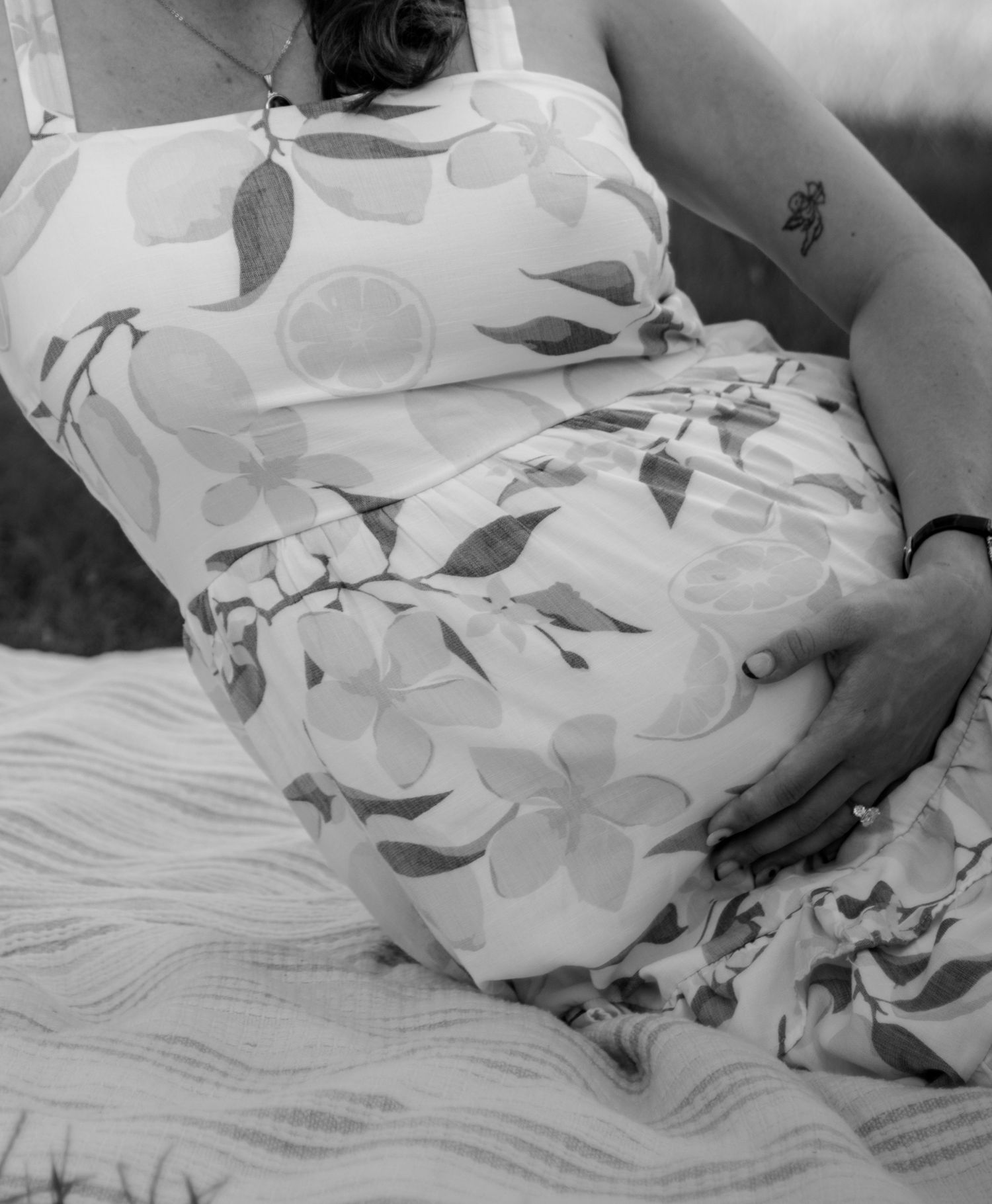Black and white photo of a pregnant woman in a floral dress cradling her baby bump outdoors.