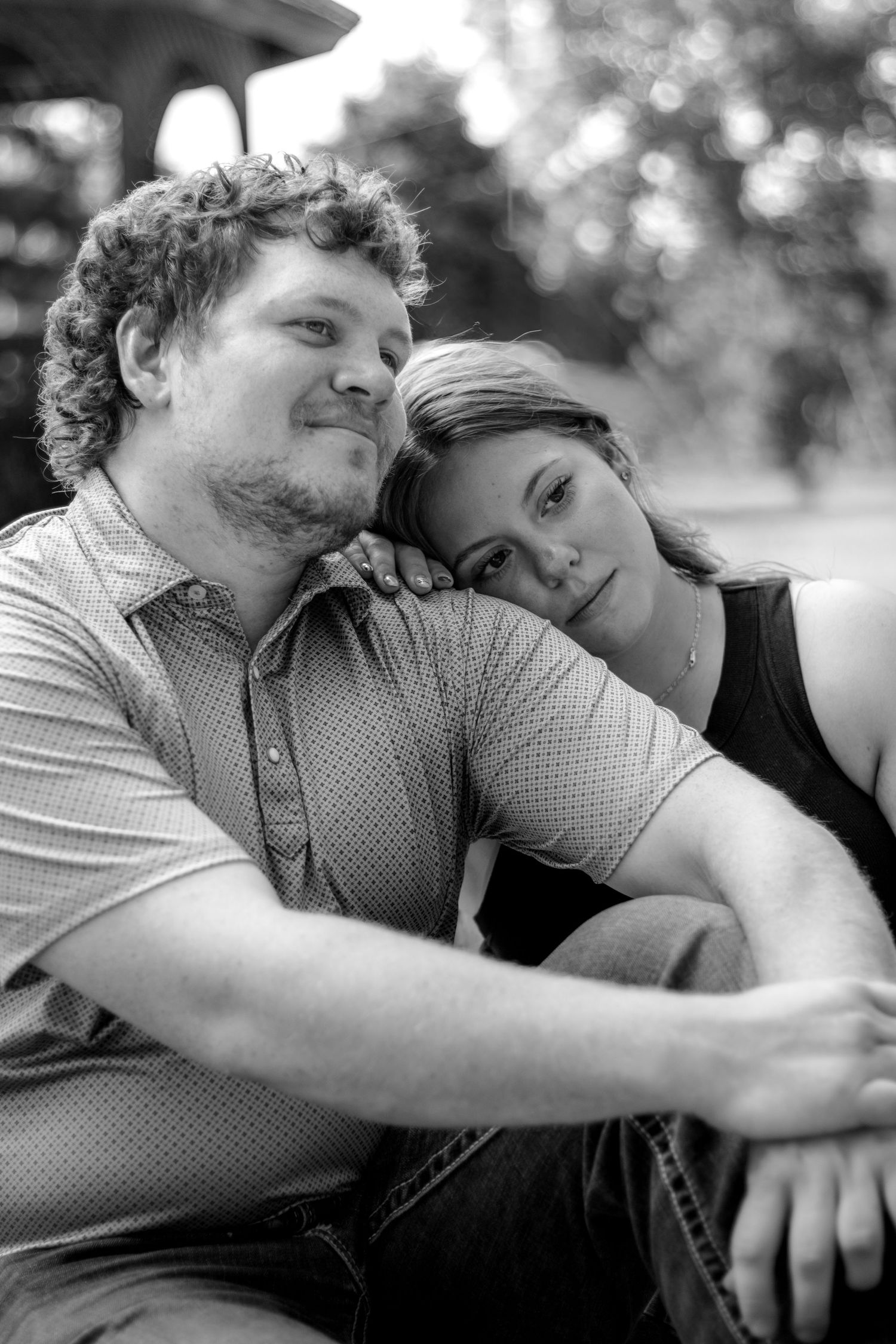 Black and white photo of a couple sitting outdoors, woman resting her head on mans shoulder affectionately.