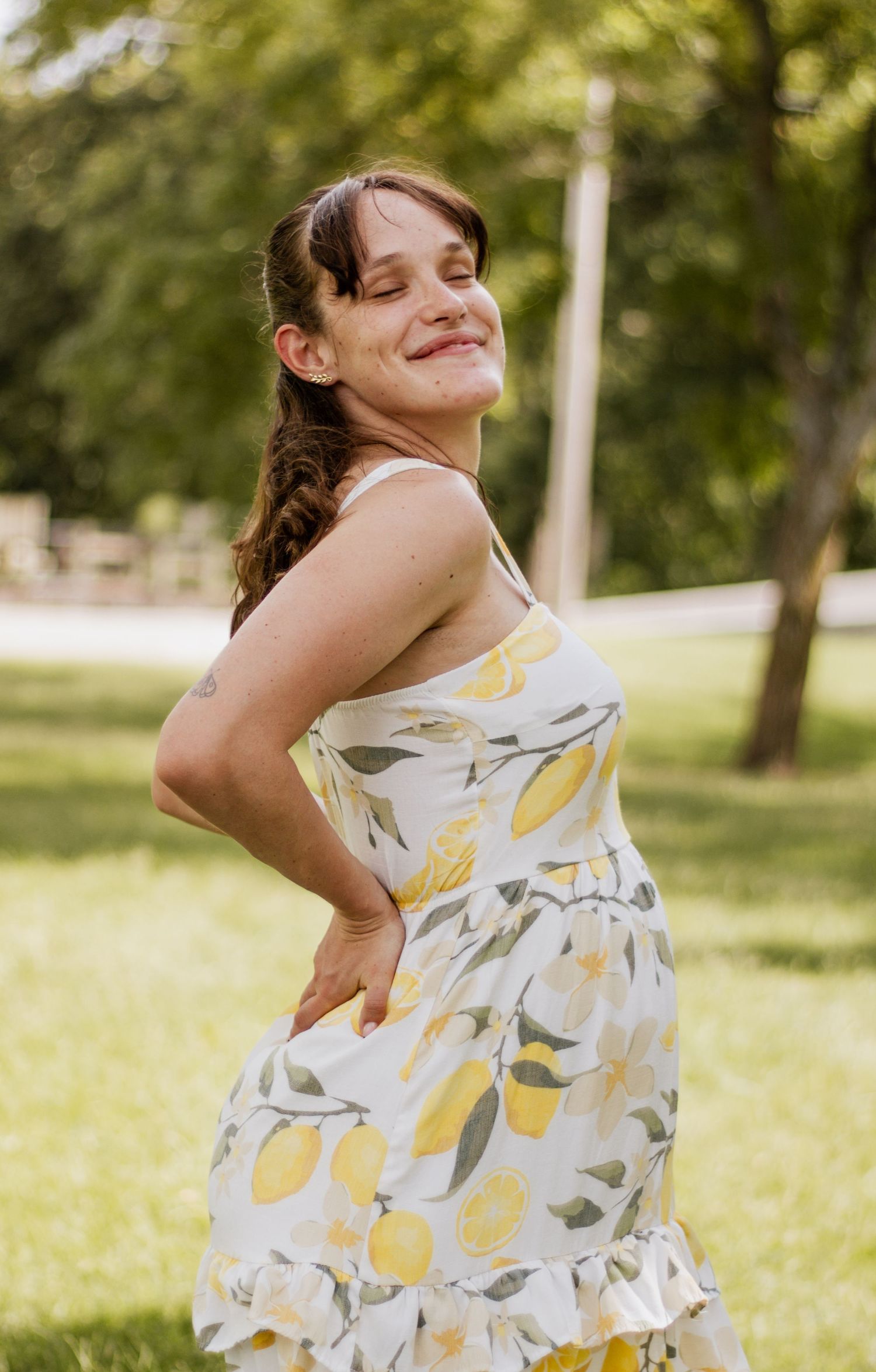 Smiling woman in a white lemon-print sundress posing outdoors in a lush green park on a sunny day.
