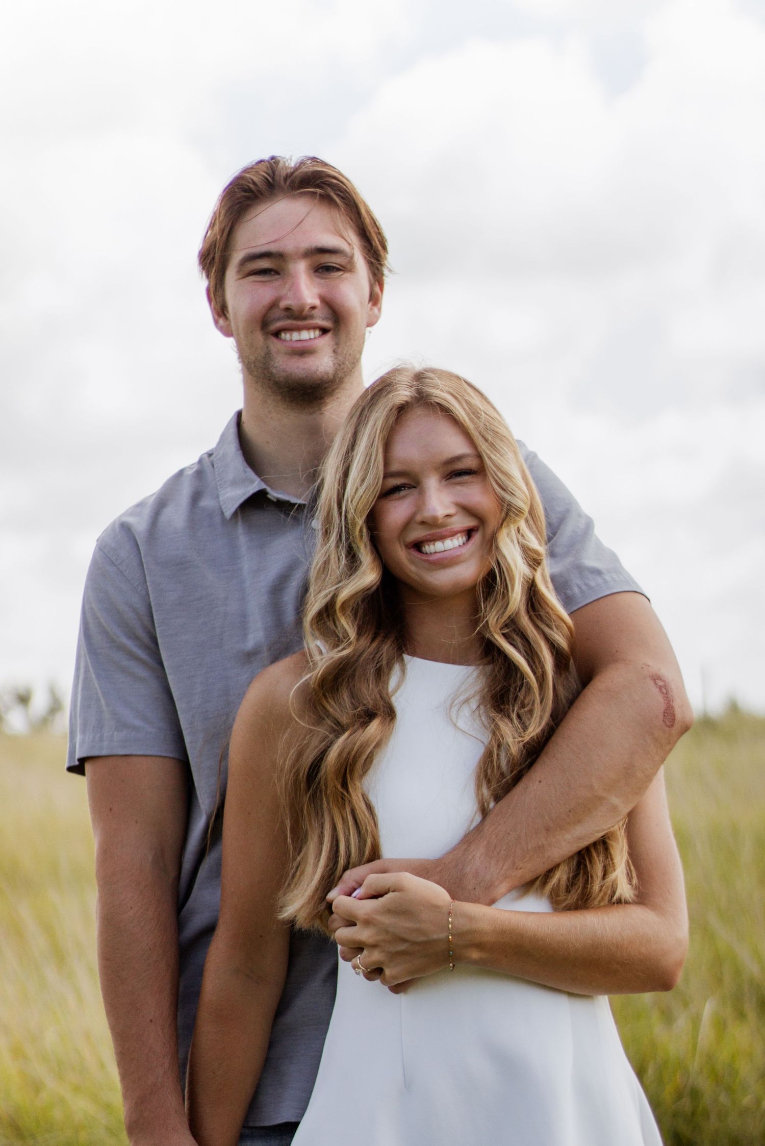 A smiling couple poses outdoors in a grassy field, the woman in white dress embracing the man in a gray polo shirt.