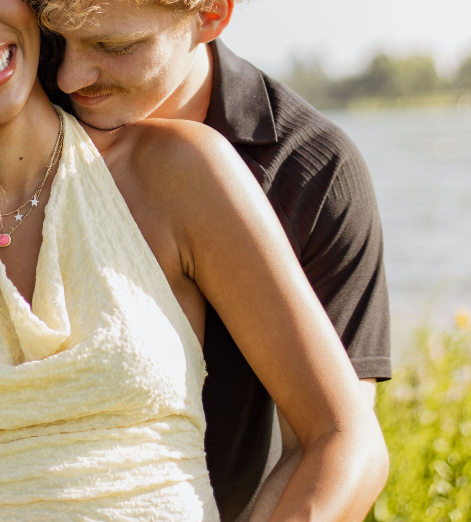 A couple embraces outdoors near a lake, woman in yellow dress with necklace, man in dark polo shirt.