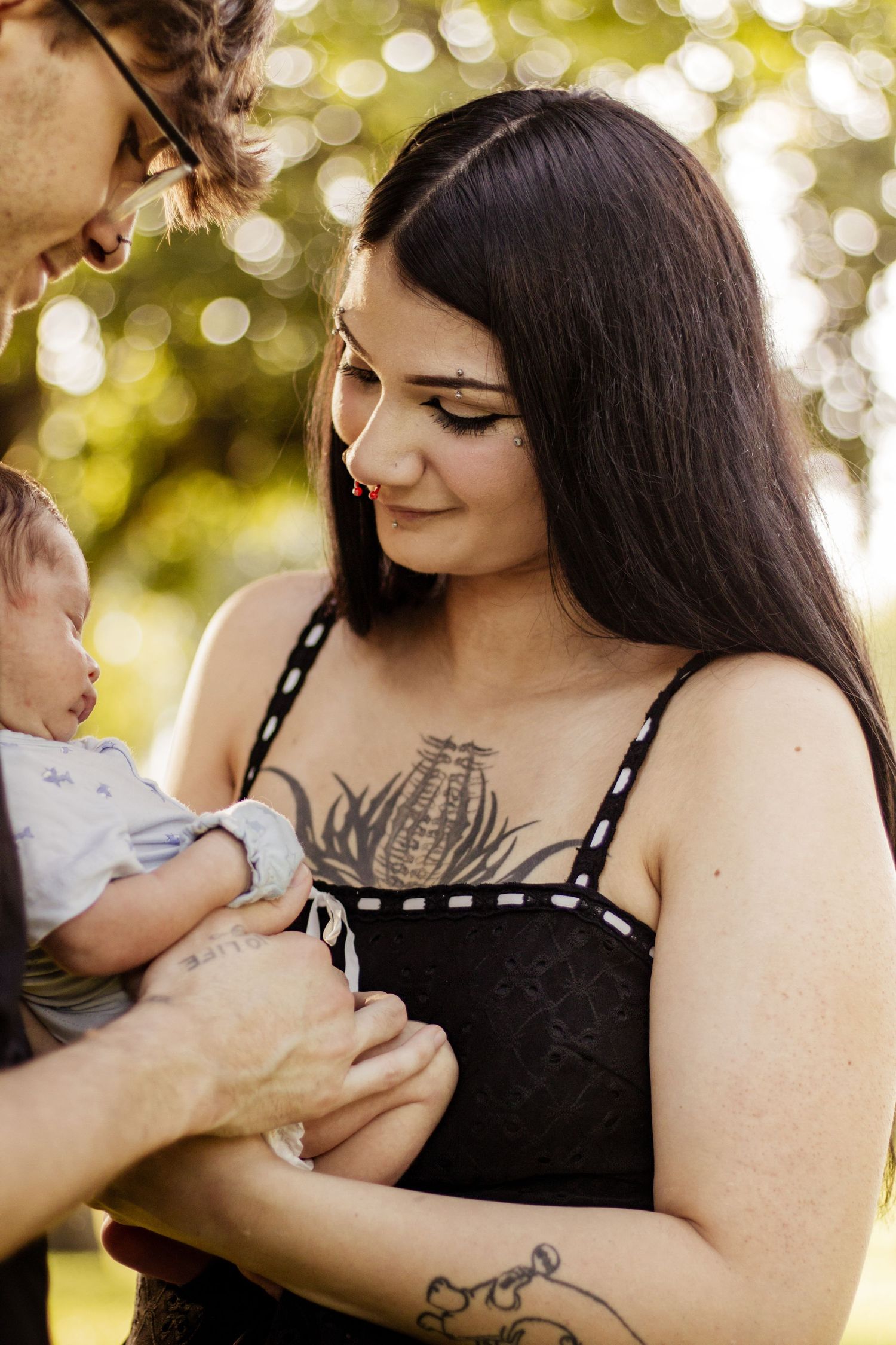 A tattooed young woman and man lovingly hold a newborn baby outdoors in a sunlit park setting.
