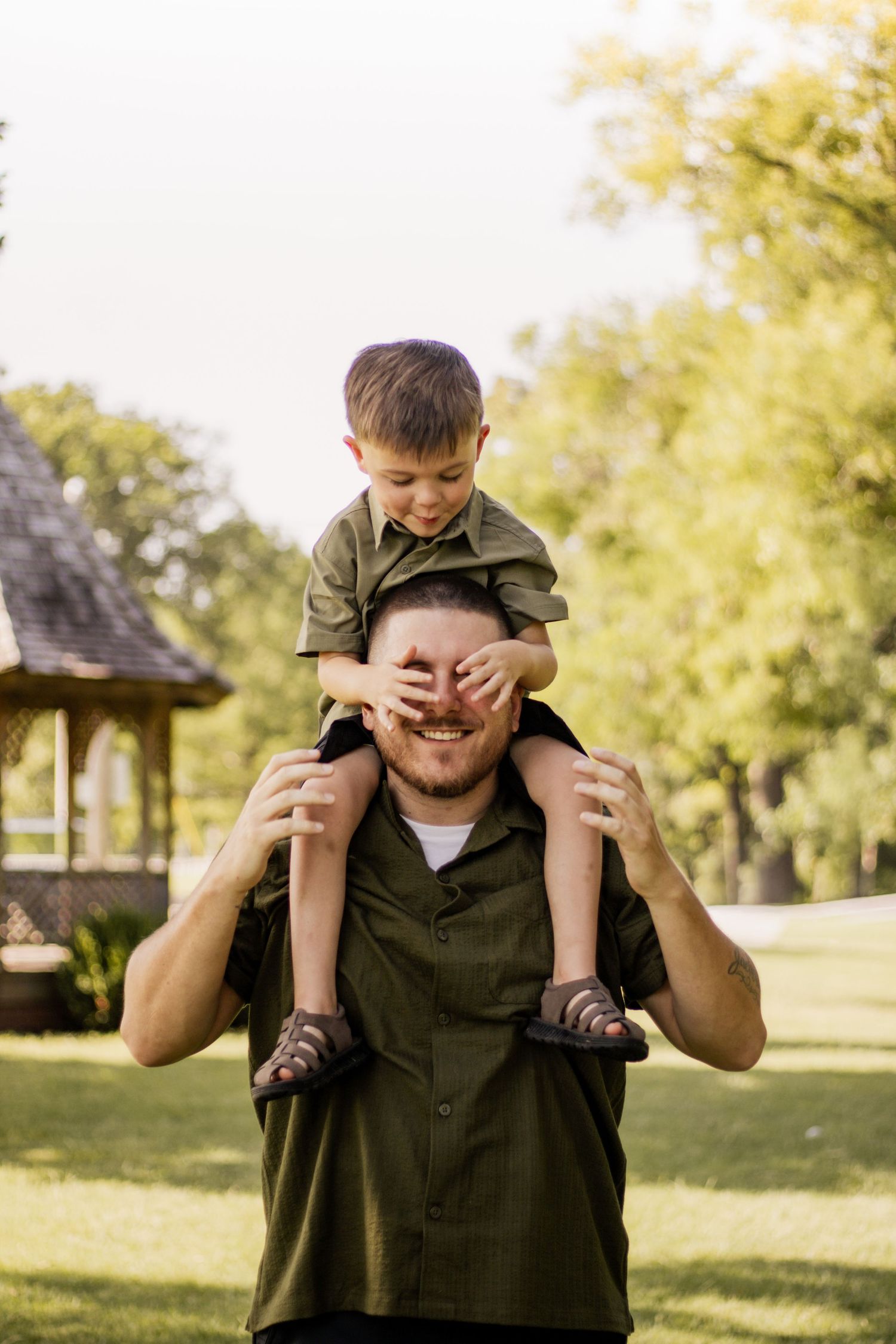 A young boy sits on his father's shoulders in a park, covering dad's eyes while both smile joyfully.