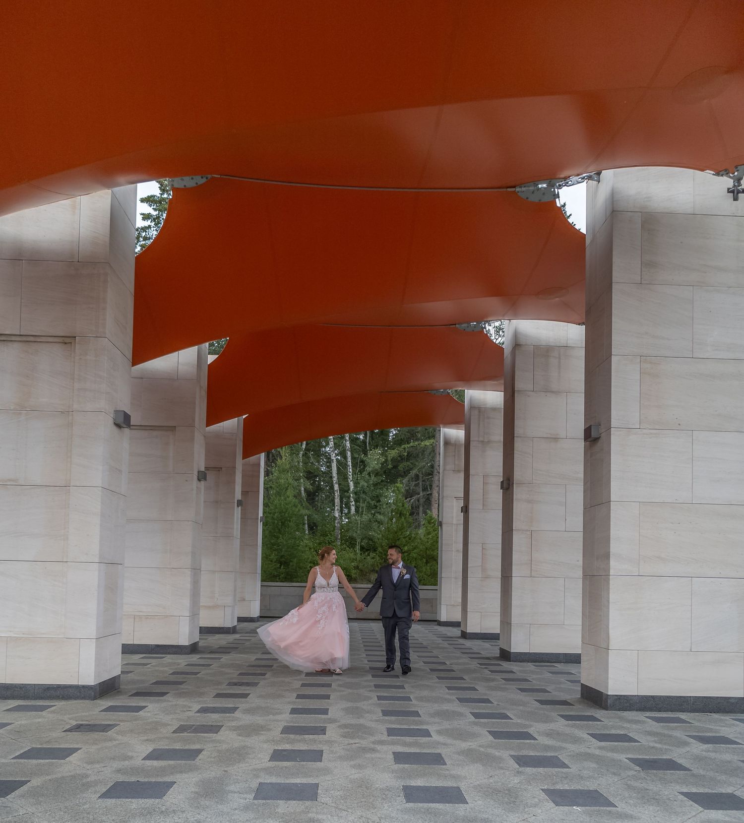 Bride and groom walk hand in hand beneath dramatic red architectural canopy.