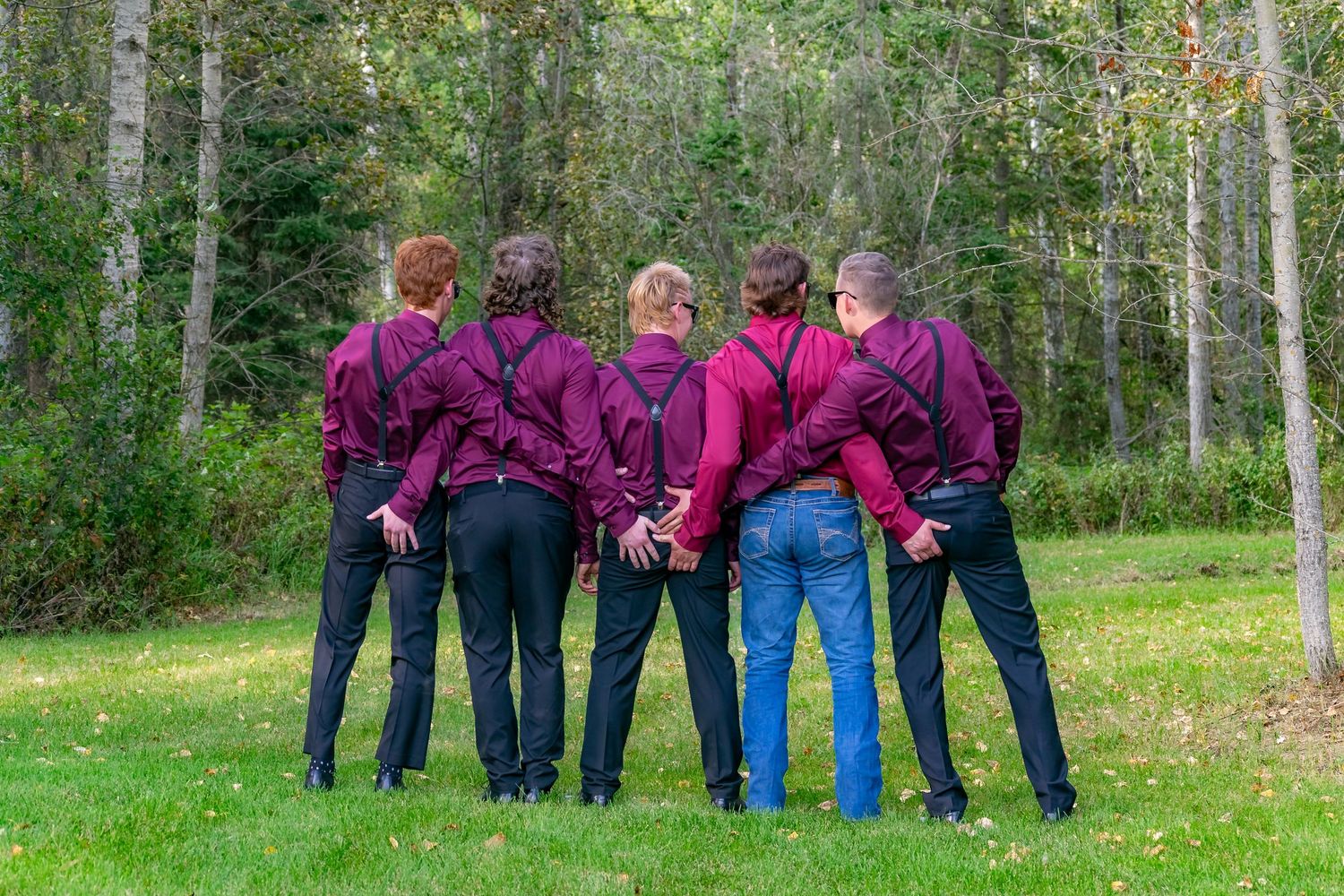 Five groomsmen in burgundy shirts standing back to back on green lawn near trees.