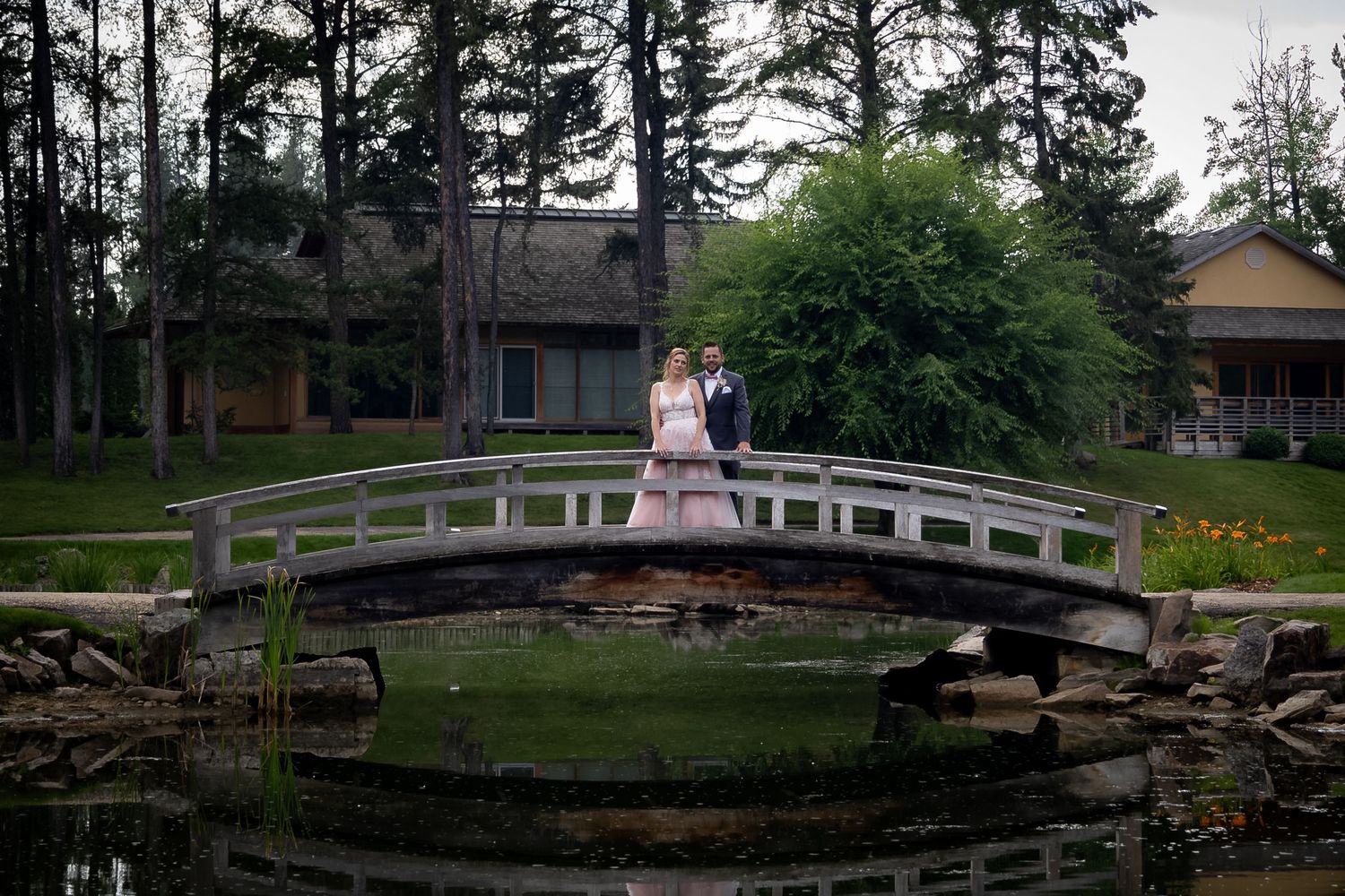Bride and groom stand on a wooden arched bridge over a pond near a lodge.