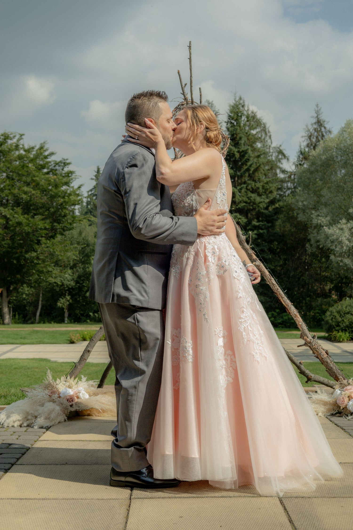 Bride and groom share first kiss outdoors in pink ballgown with rustic branch arch backdrop.