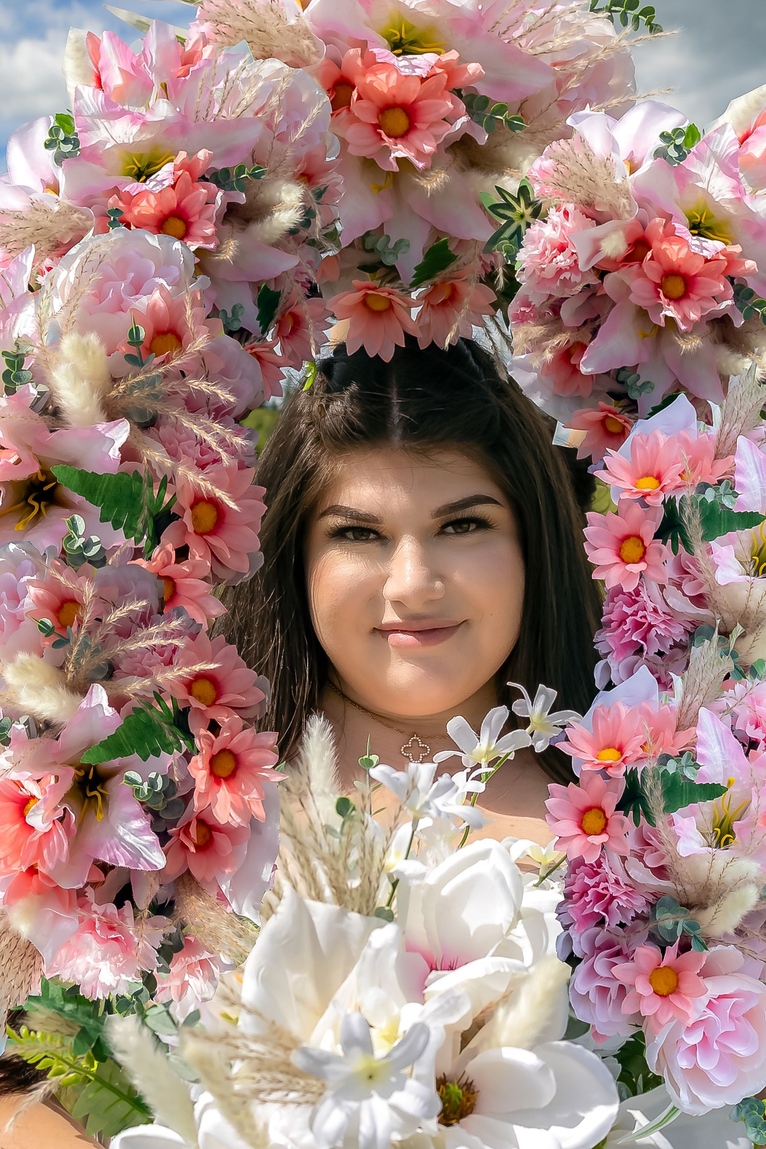 Young woman framed by beautiful pink and white flower wreath holding white bouquet outdoors.