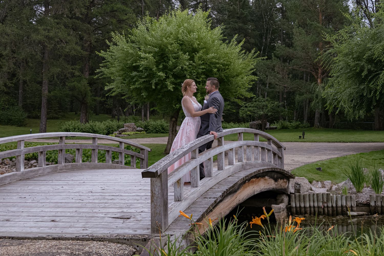 Bride and groom share a romantic moment on a wooden arched bridge surrounded by lush green trees in a garden park.