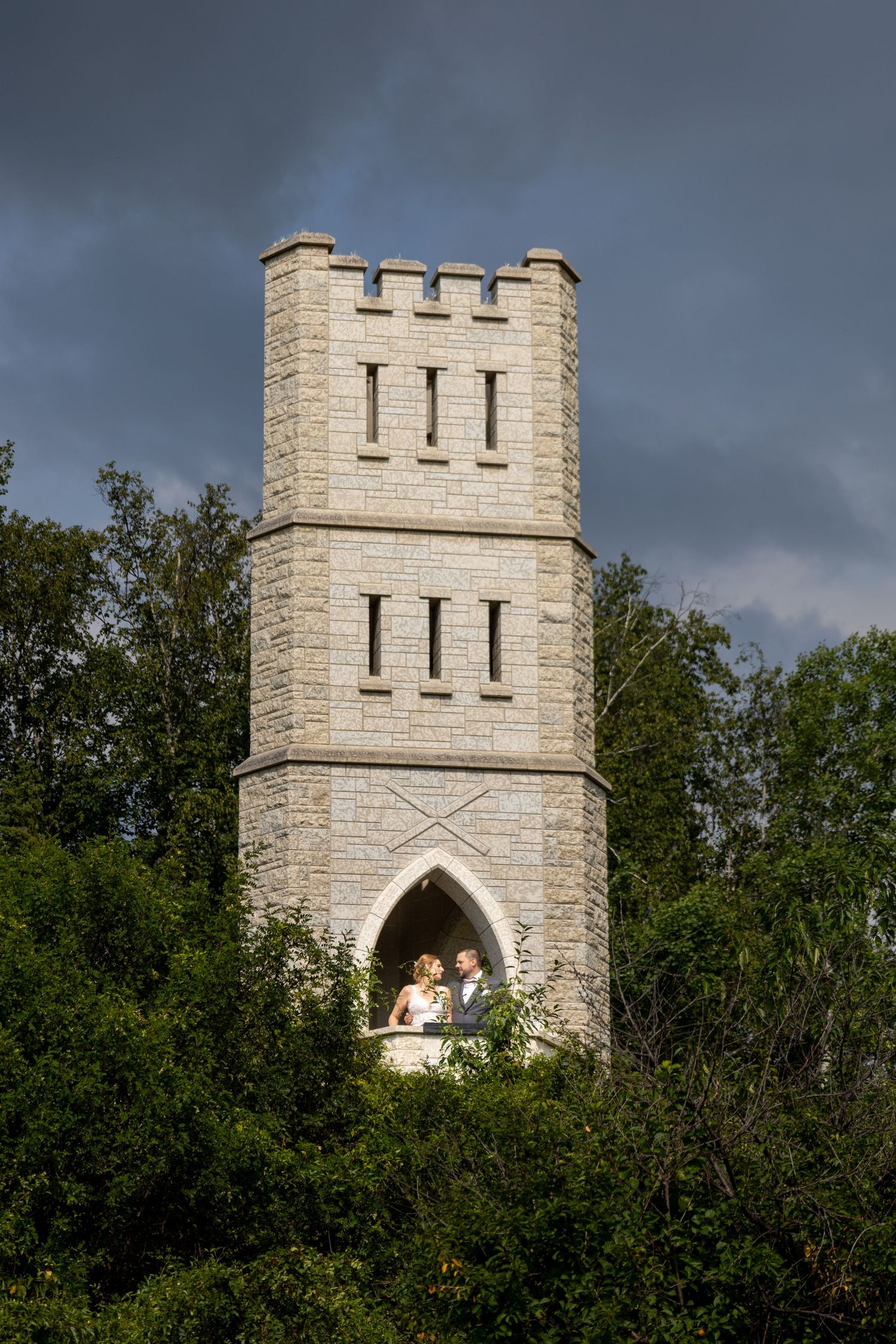 Bride and groom pose in arched window of stone castle tower surrounded by lush trees.