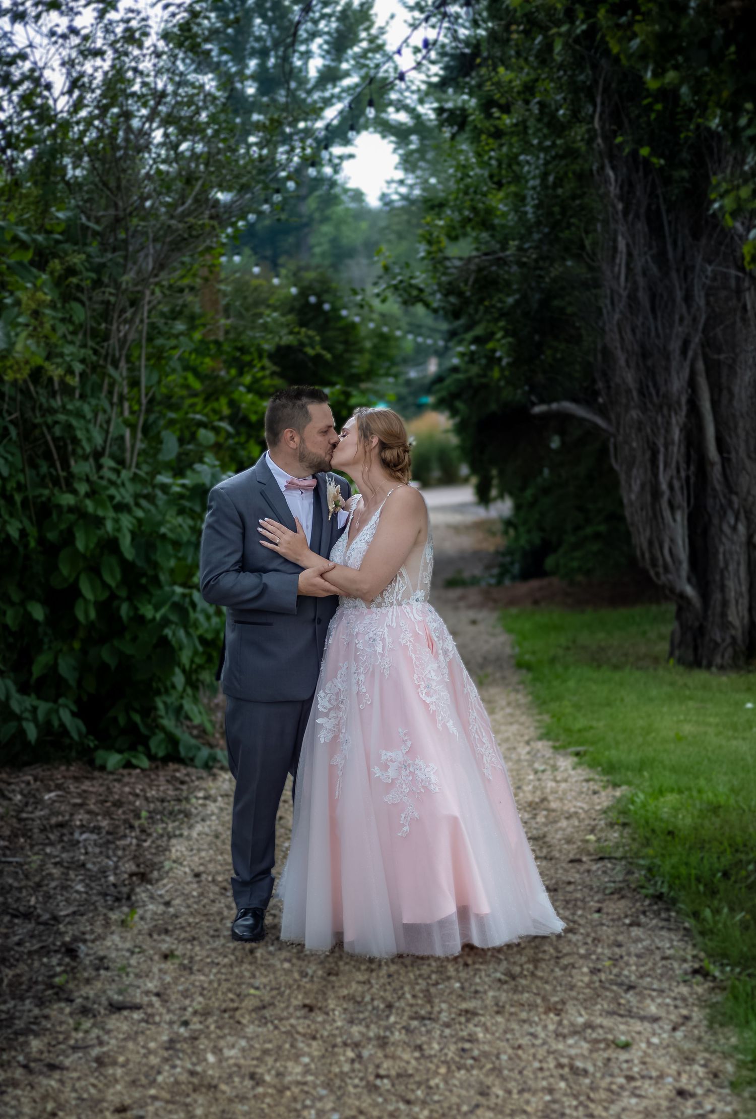 Wedding couple shares a kiss on a tree-lined gravel path, bride in pink gown.