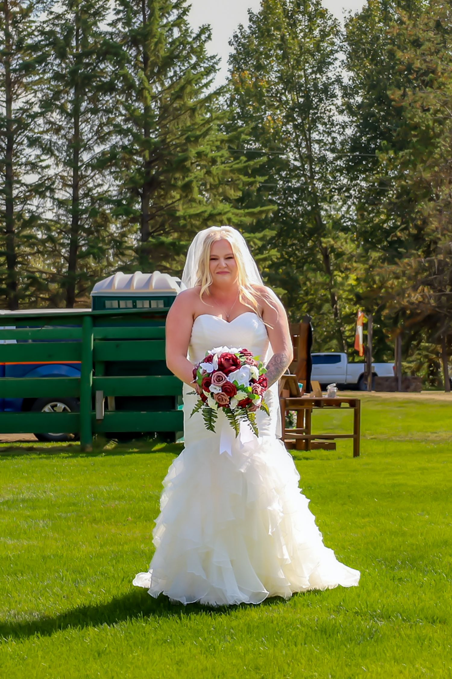 Blonde bride in white mermaid wedding dress holding red and white bouquet outdoors.