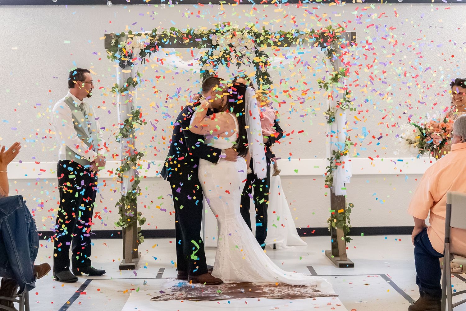 Newlyweds share first kiss under colorful confetti shower at indoor wedding ceremony with floral arch.