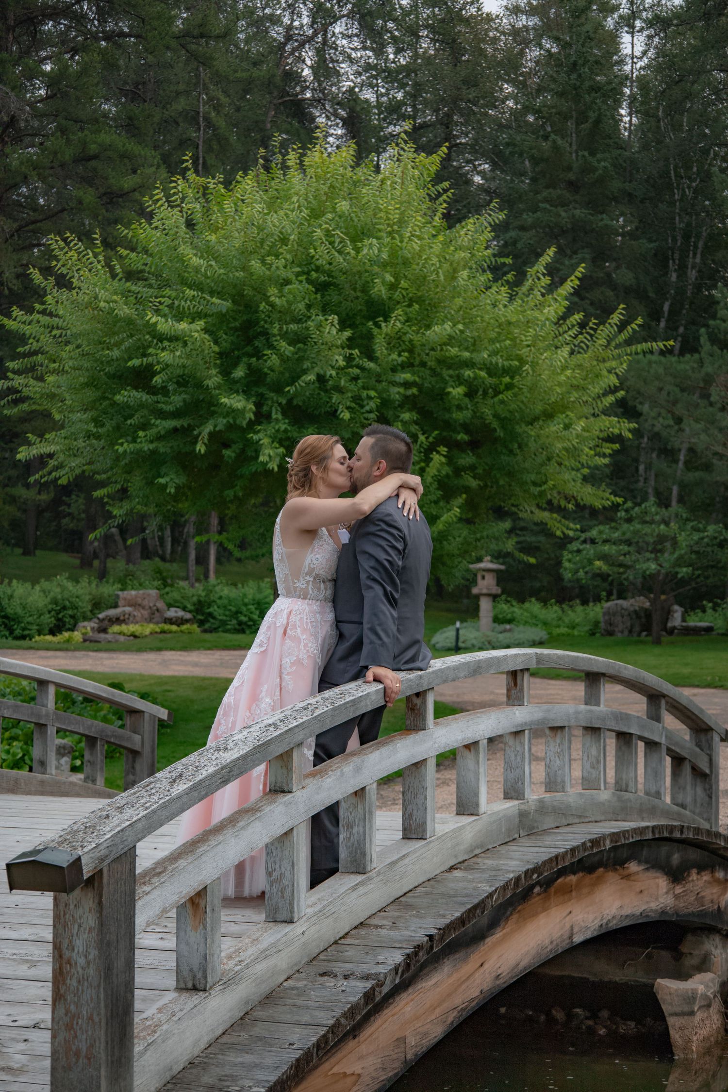 Bride and groom share a kiss on a rustic wooden arched bridge in a garden.
