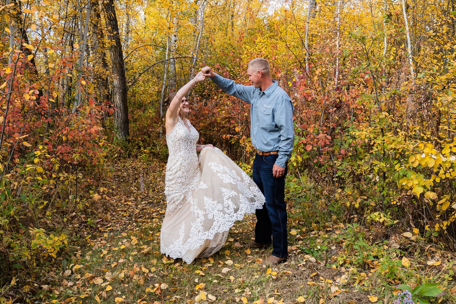 Groom twirling bride in a stunning lace gown among colorful autumn trees in a forest setting.
