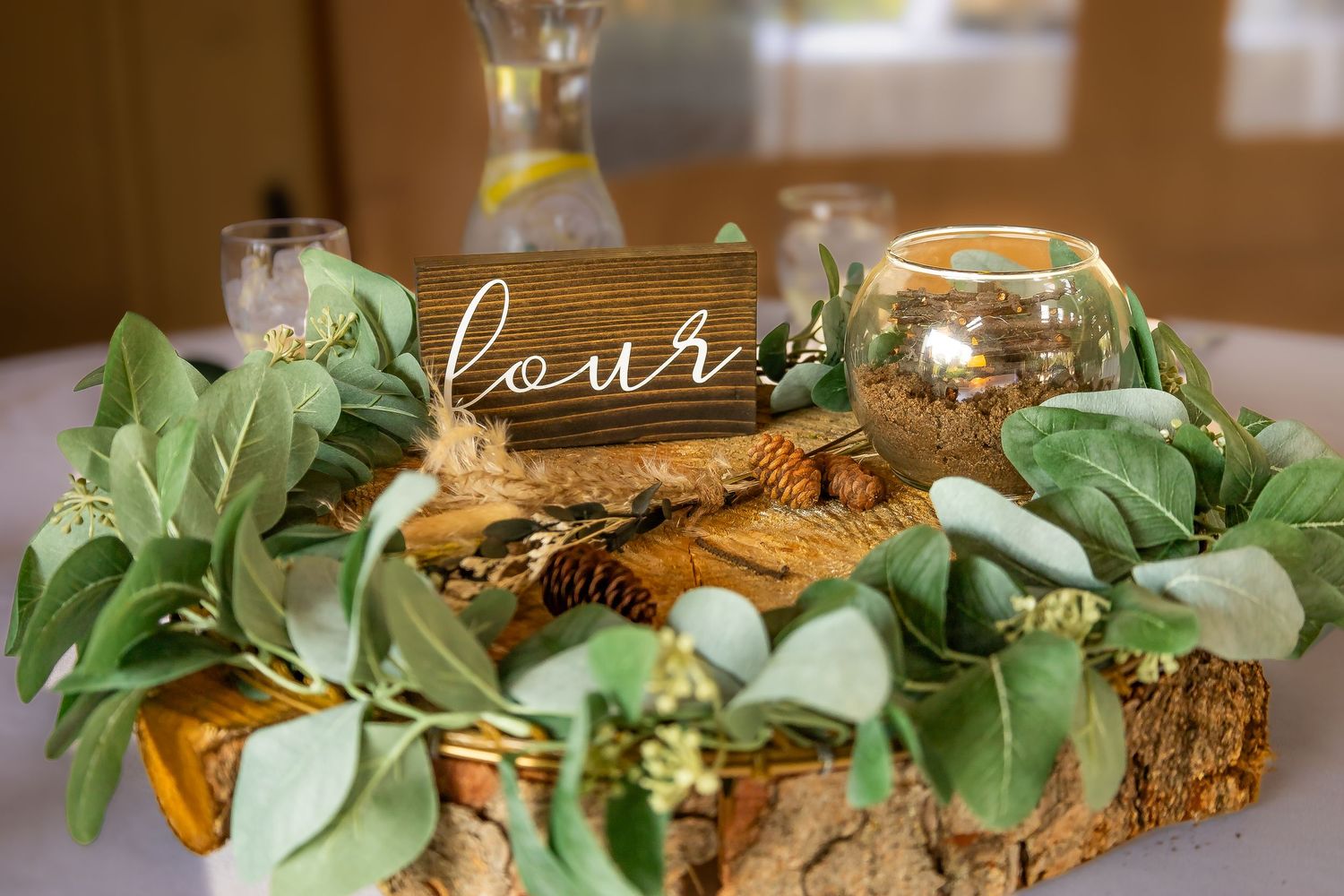 Rustic wedding table centerpiece with greenery, pinecones, candle, and wooden number four sign.