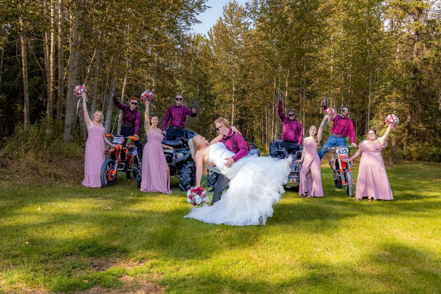 Wedding party celebrating with dirt bike and quads in a sunny forest clearing, bridesmaids in pink dresses.
