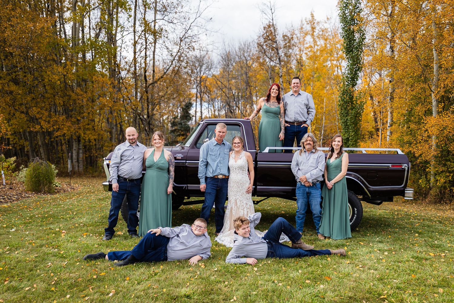 Fall wedding party group photo with bride and groom in front of a classic dark pickup truck surrounded by golden trees.