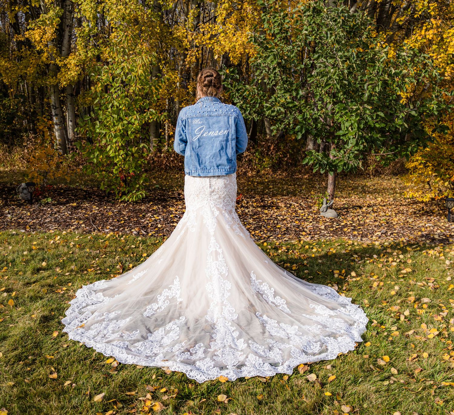 Bride in lace mermaid gown and custom denim jacket stands amid vibrant autumn trees.