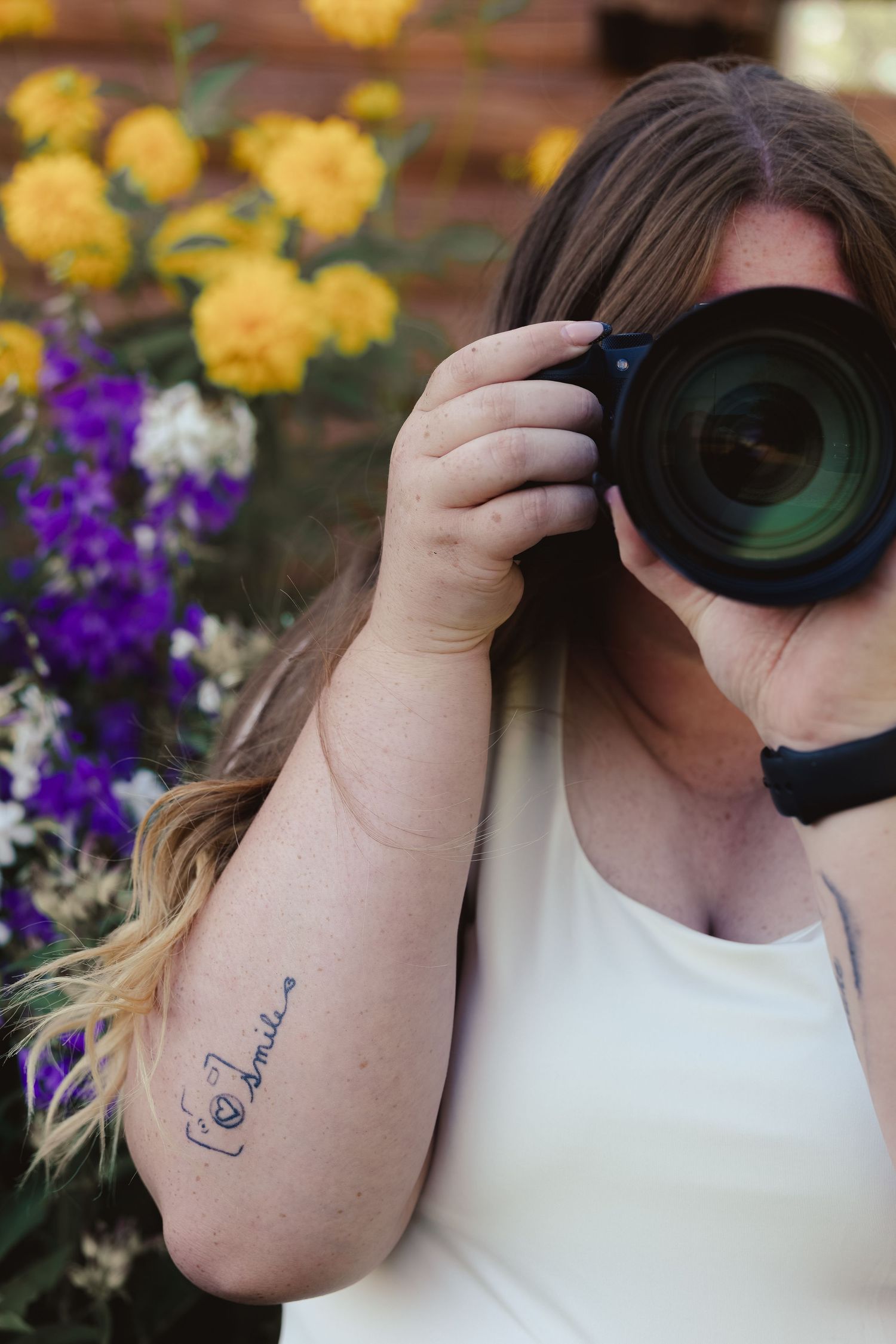 Woman with tattoo on arm holds DSLR camera up to her face surrounded by colorful yellow and purple flowers.