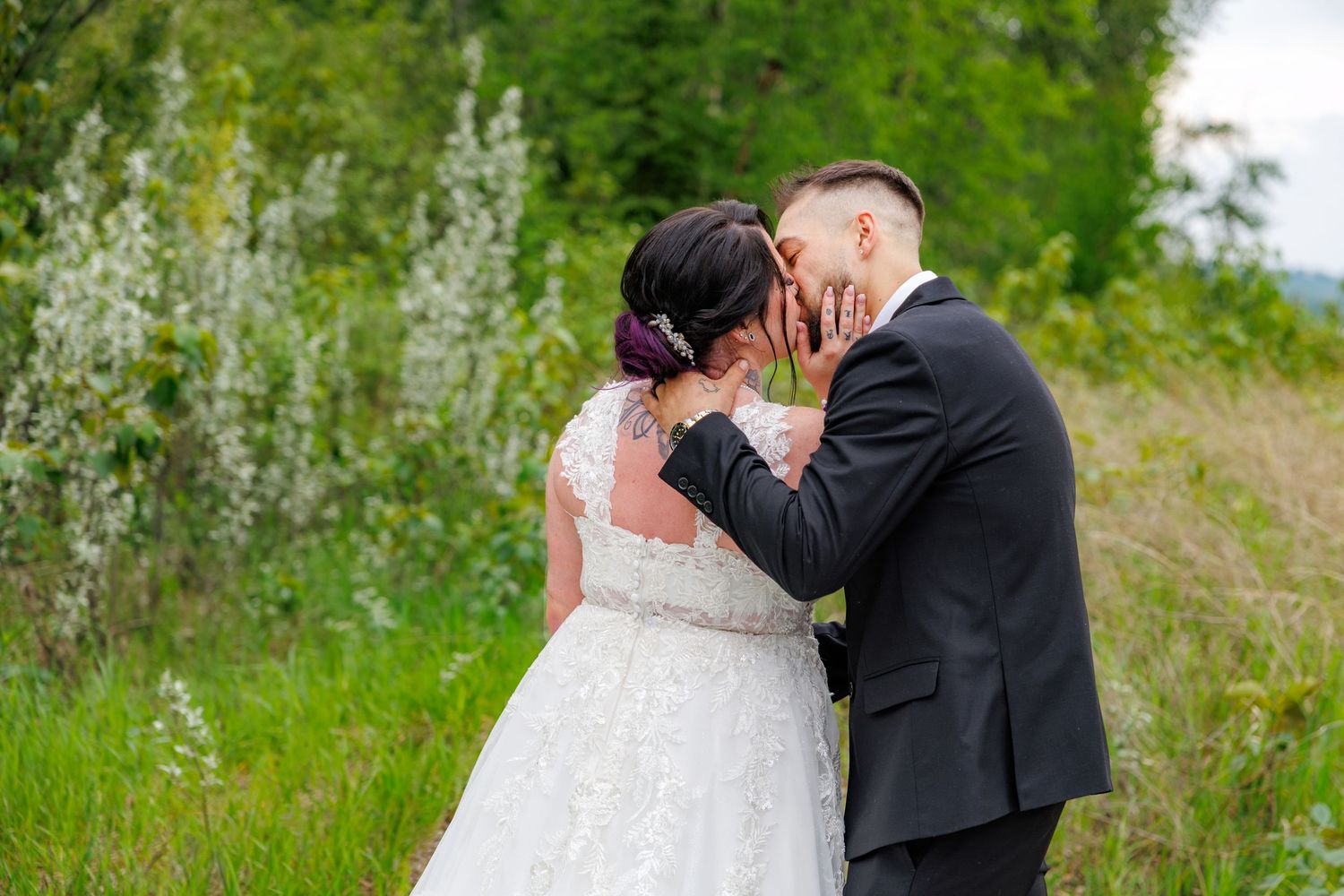 Bride and groom share an intimate kiss in a green meadow, bride in lace gown with veil.