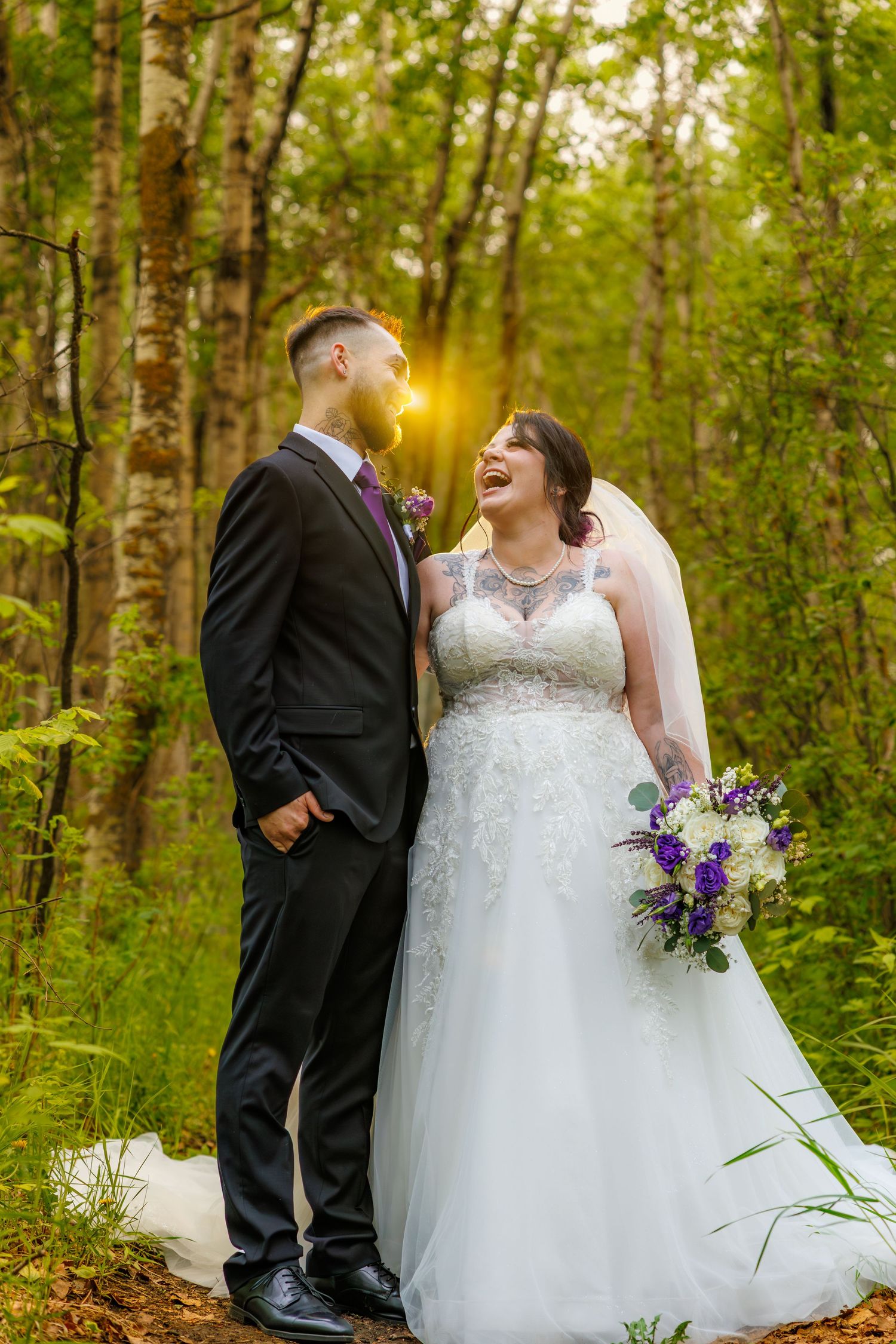 Laughing bride in lace gown holds purple bouquet beside suited groom in sunlit forest wedding portrait.