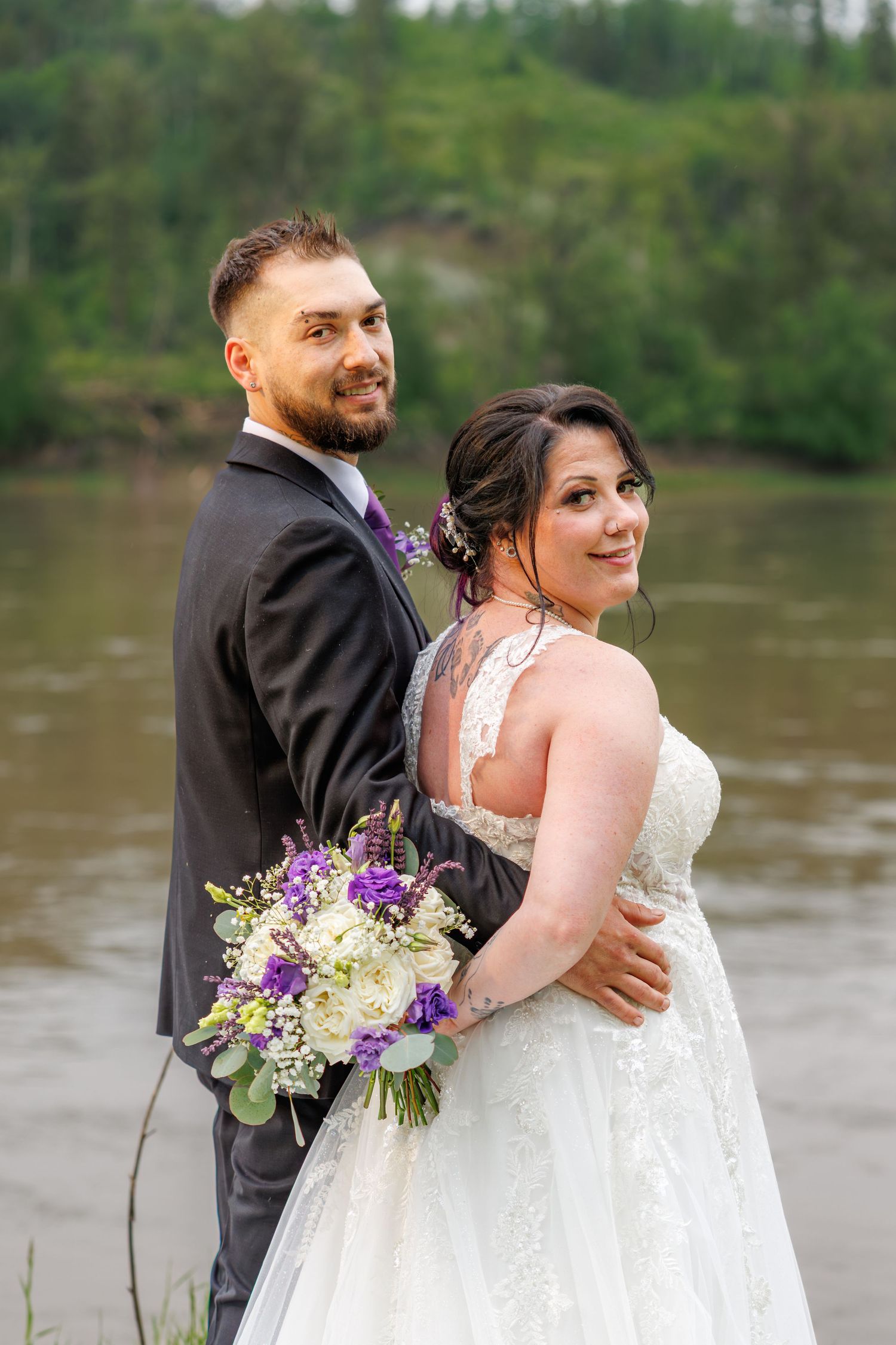 Smiling bride and groom pose together near a river, bride holding purple and white floral bouquet.