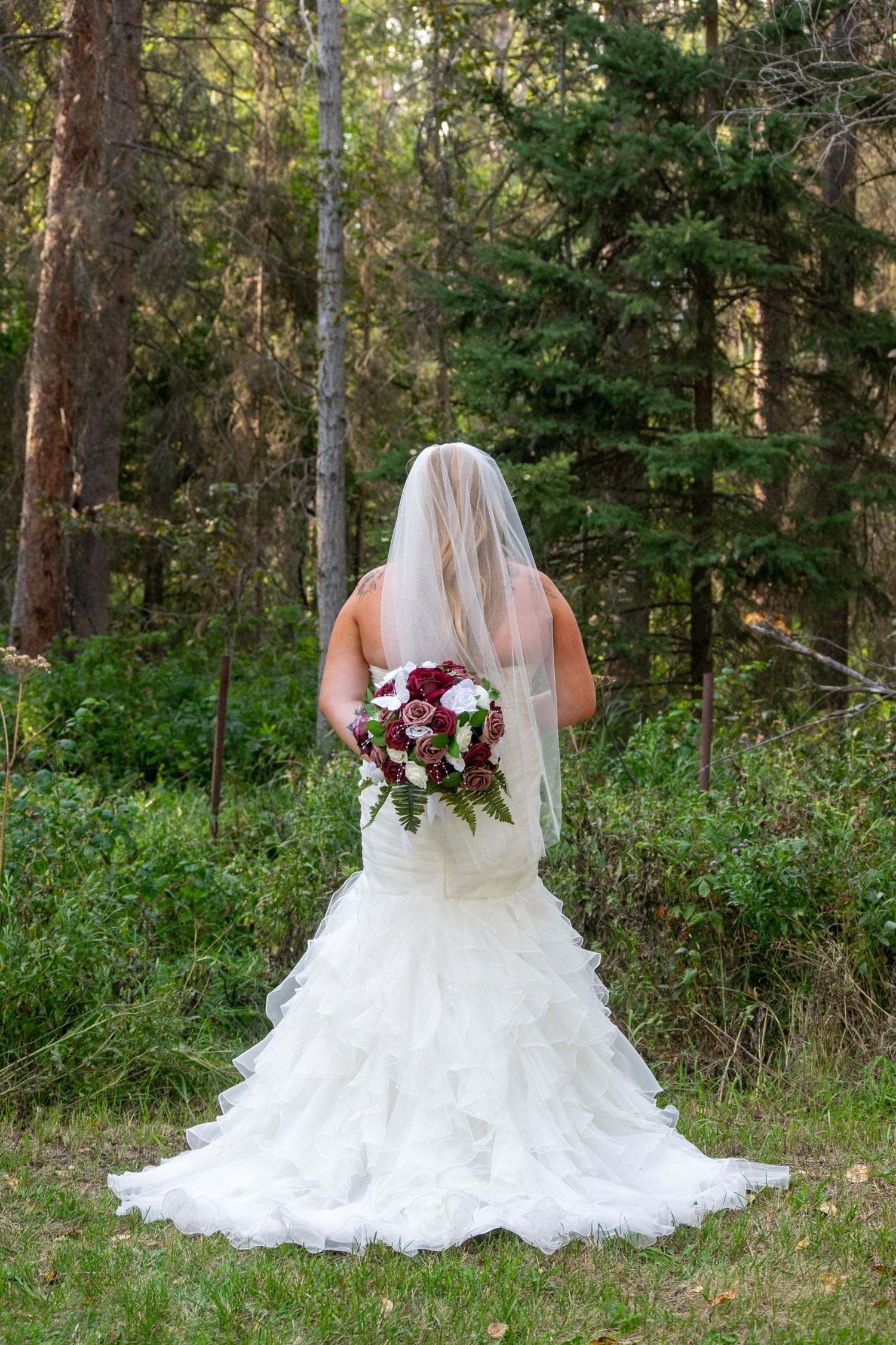 Bride in white mermaid wedding dress with veil holding a red and white bouquet in a forest.